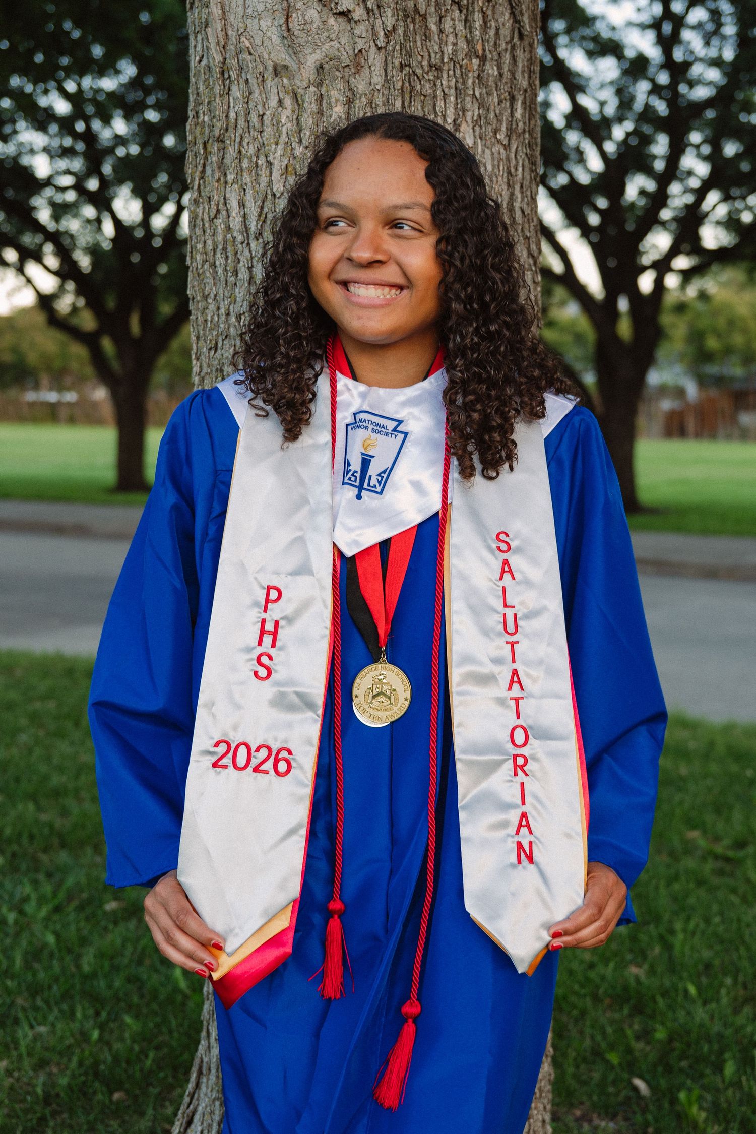 Senior in her grad gown and awards smiling to the side in a large tree-filled park in Richardson, Texas for a senior photo session