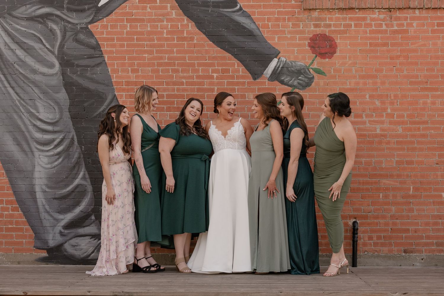 Bride and her wedding party wearing dresses in shades of green and pink poses against a brick wall with dramatic mural artwork.
