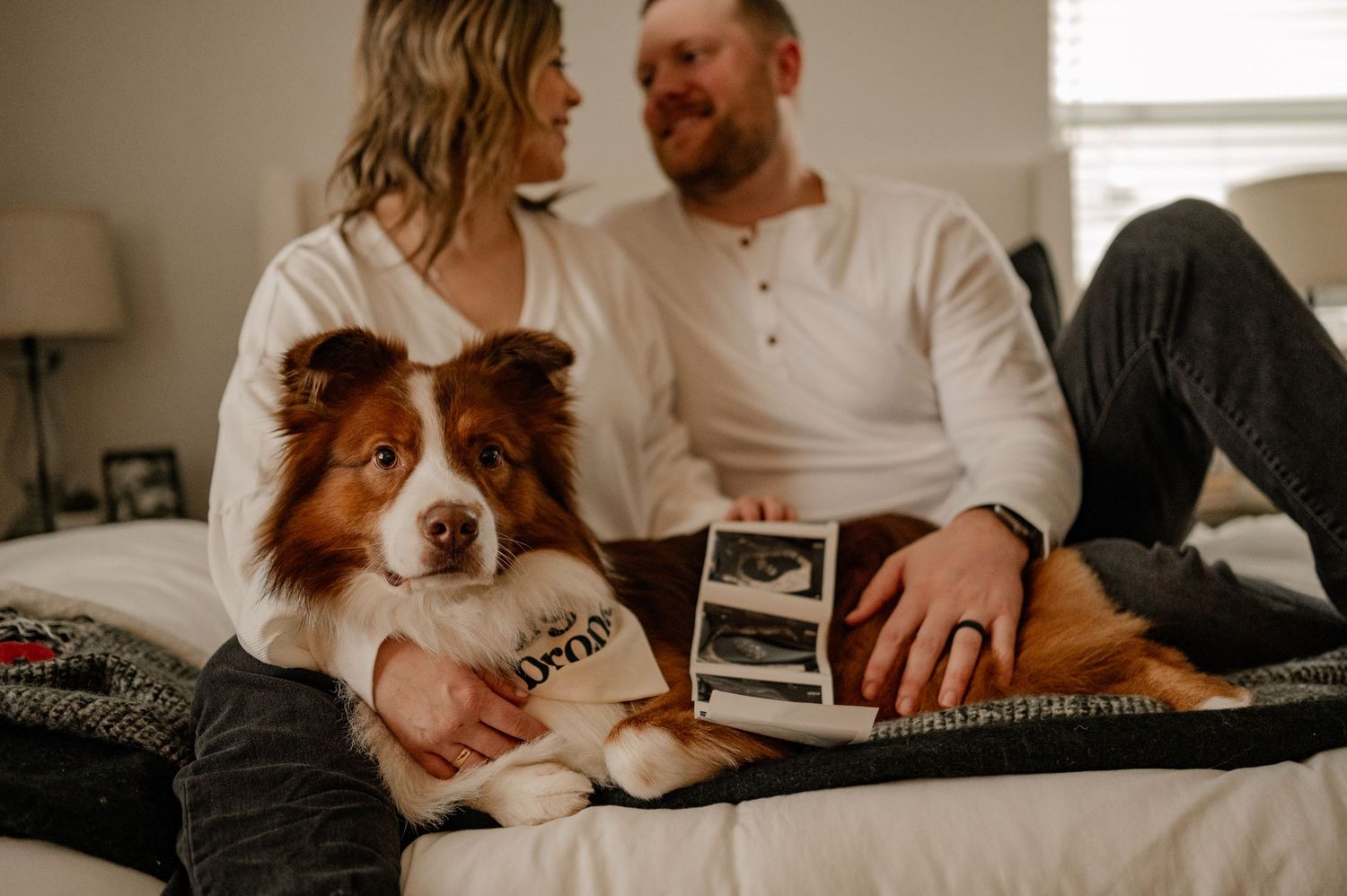 Couple relaxing on bed with their Australian Shepherd dog while looking at ultrasound photos.