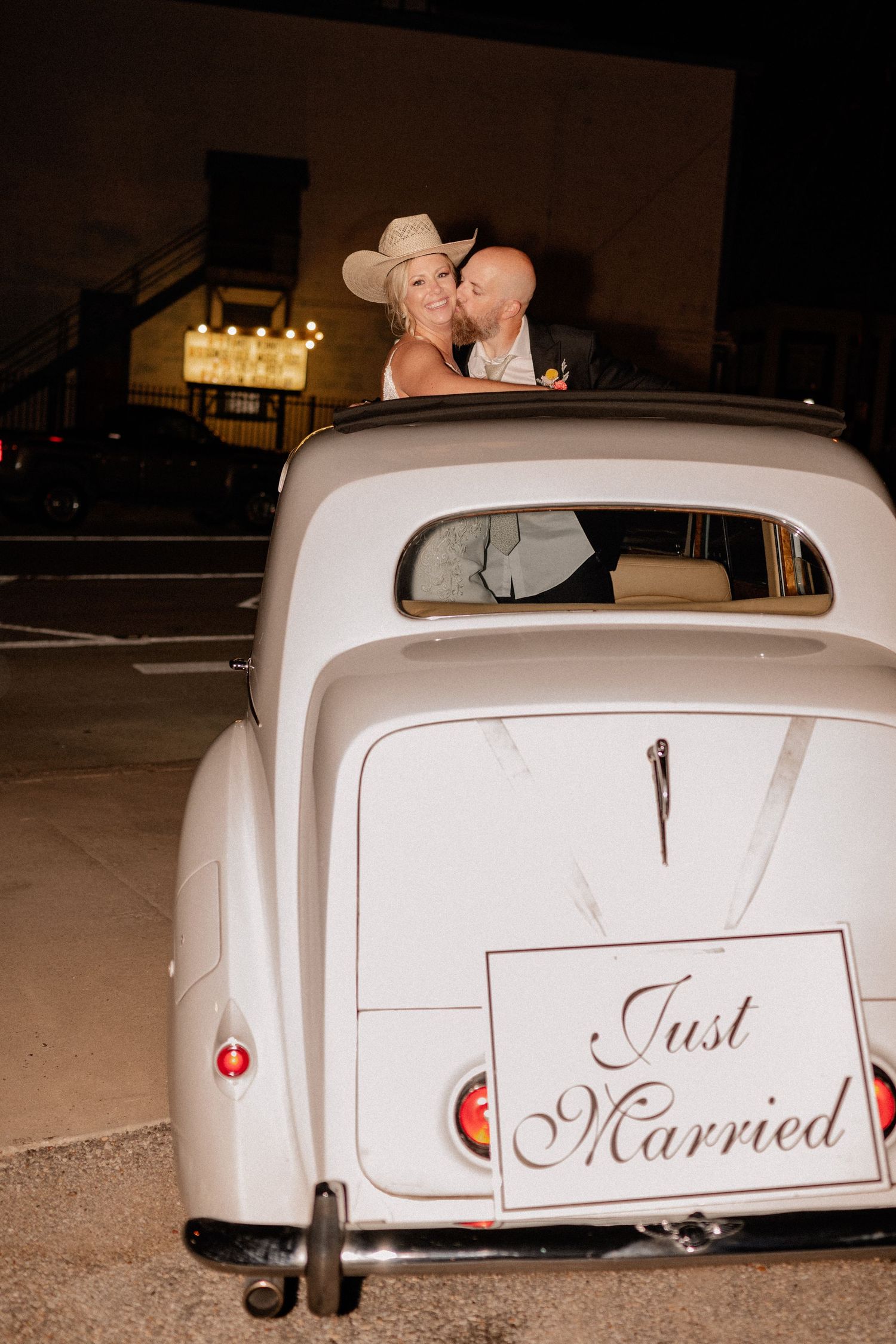Flash photo of bride and groom exiting the wedding reception in a vintage white car with a Just Married sign