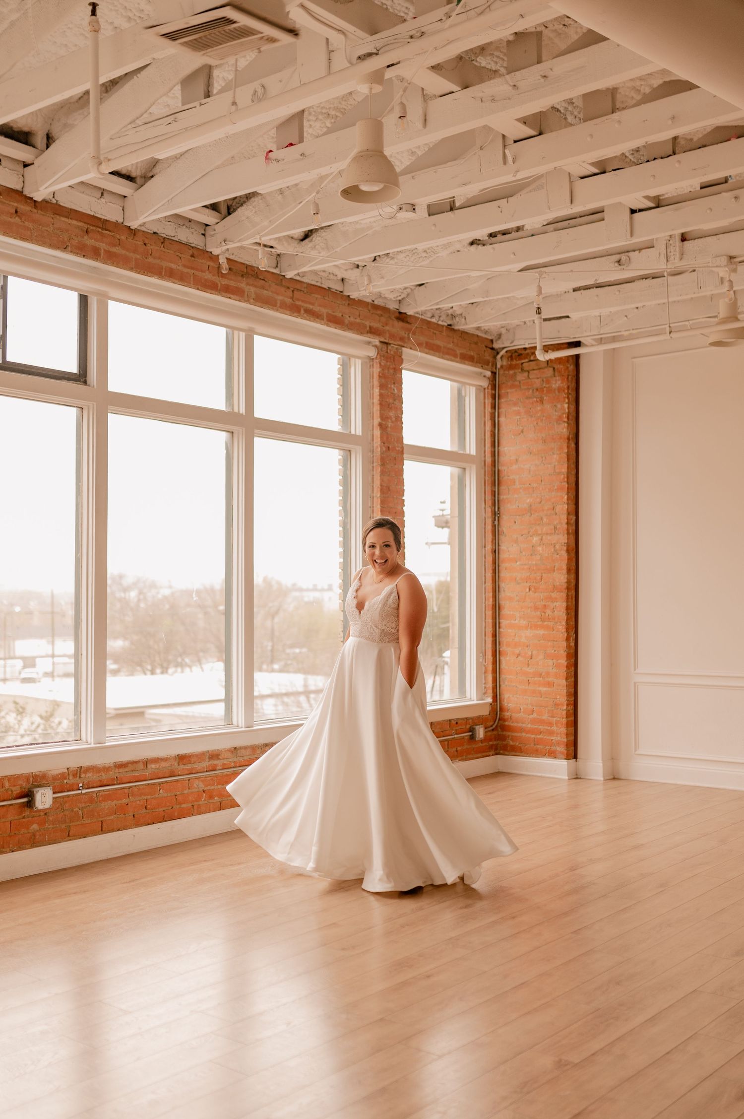Bride in flowing white dress poses in sunlit industrial space with exposed brick and large windows.