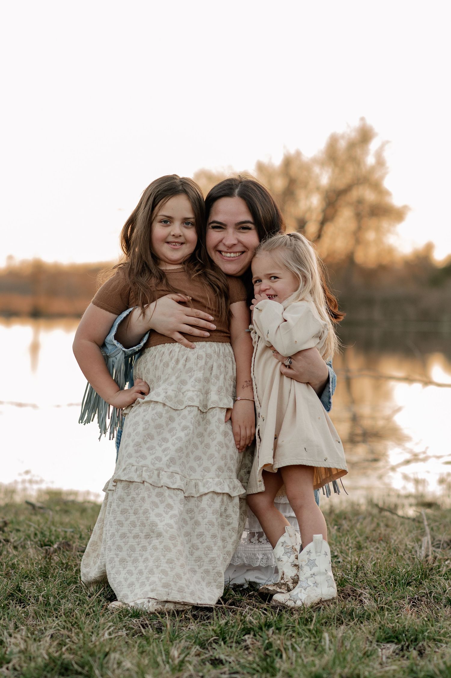 Mother hugging her daughters in front of a large pond on their Rockwall Texas farm for a fall at-home family photo session at golden hour