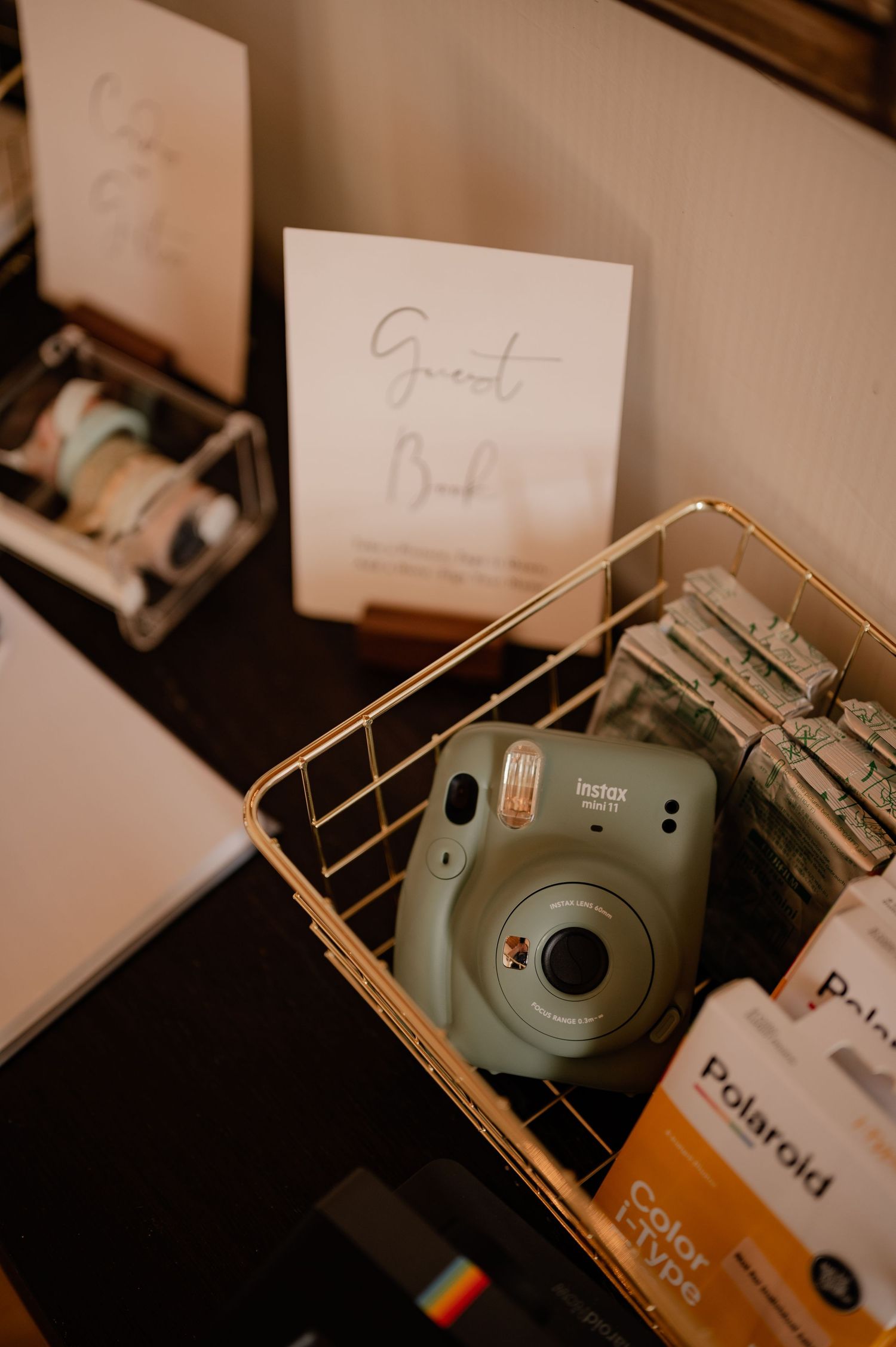 A mint green Mini instant polaroid camera sits in a basket with Polaroid film and papers for a custom wedding guest book