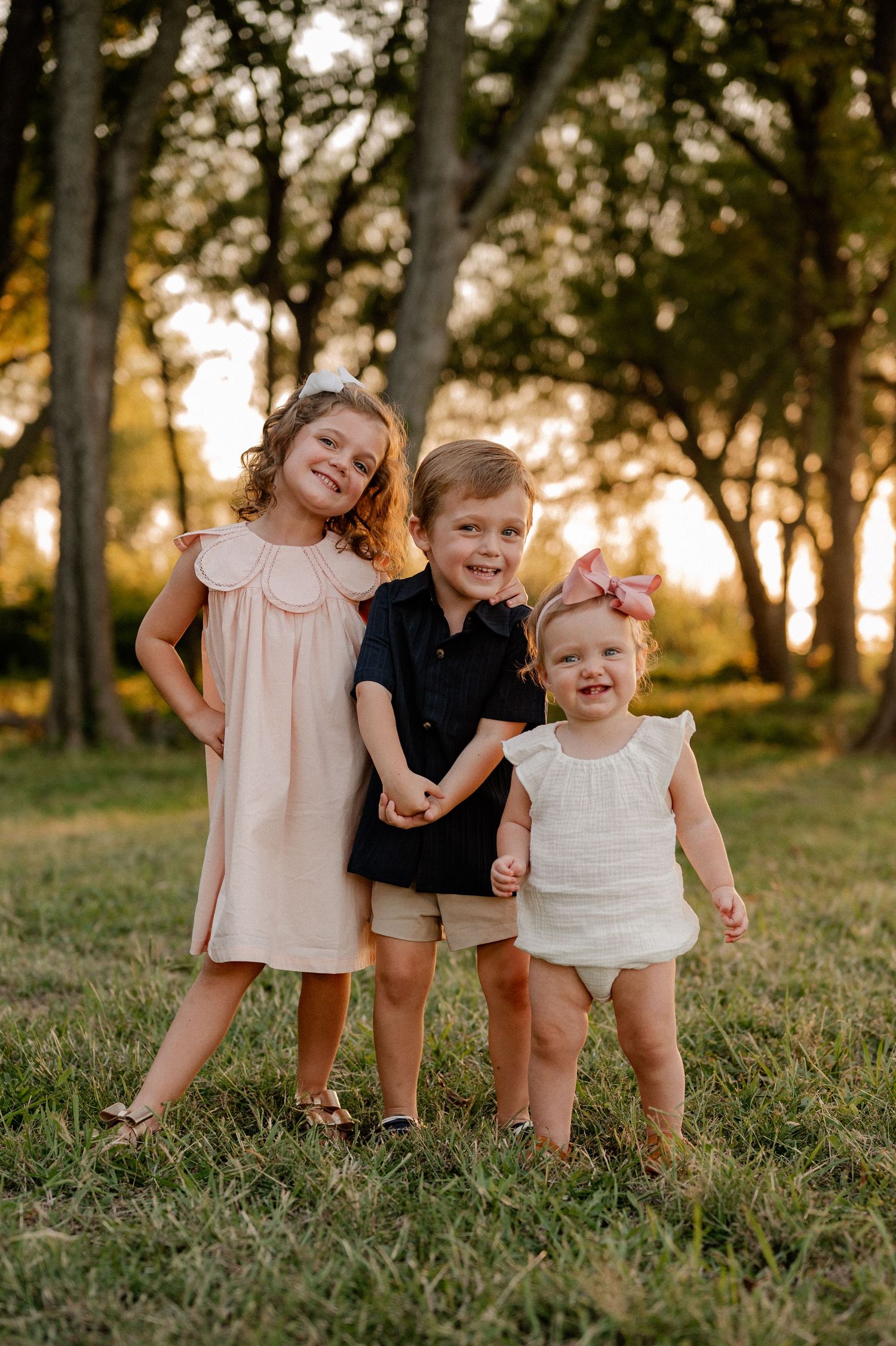 Three siblings posing with big smiles for Summer family photos in a treelined field with the sun setting over the lake in Rockwall, Texas