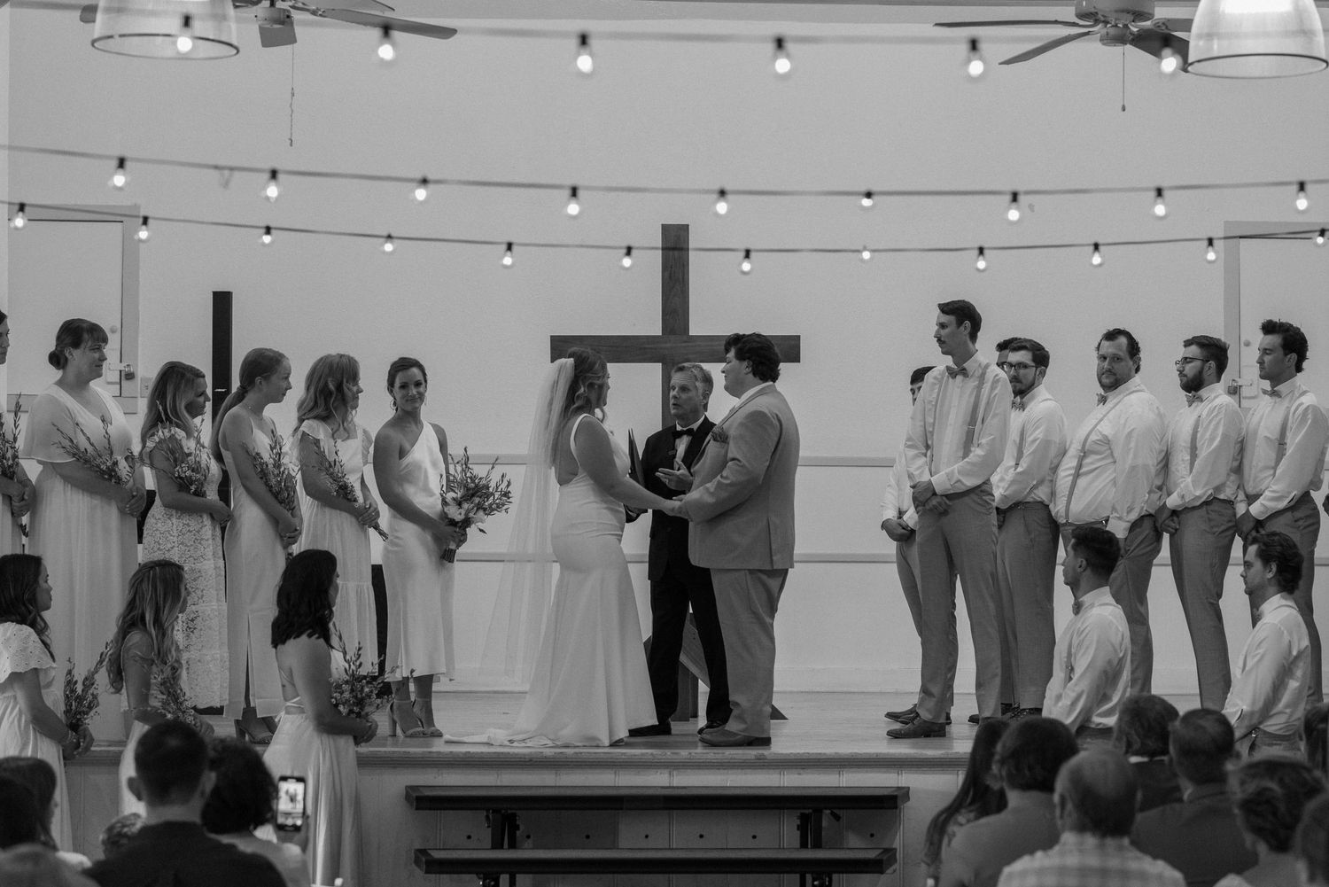 Black and white photo of a wedding ceremony in a bright indoor venue with string lights.