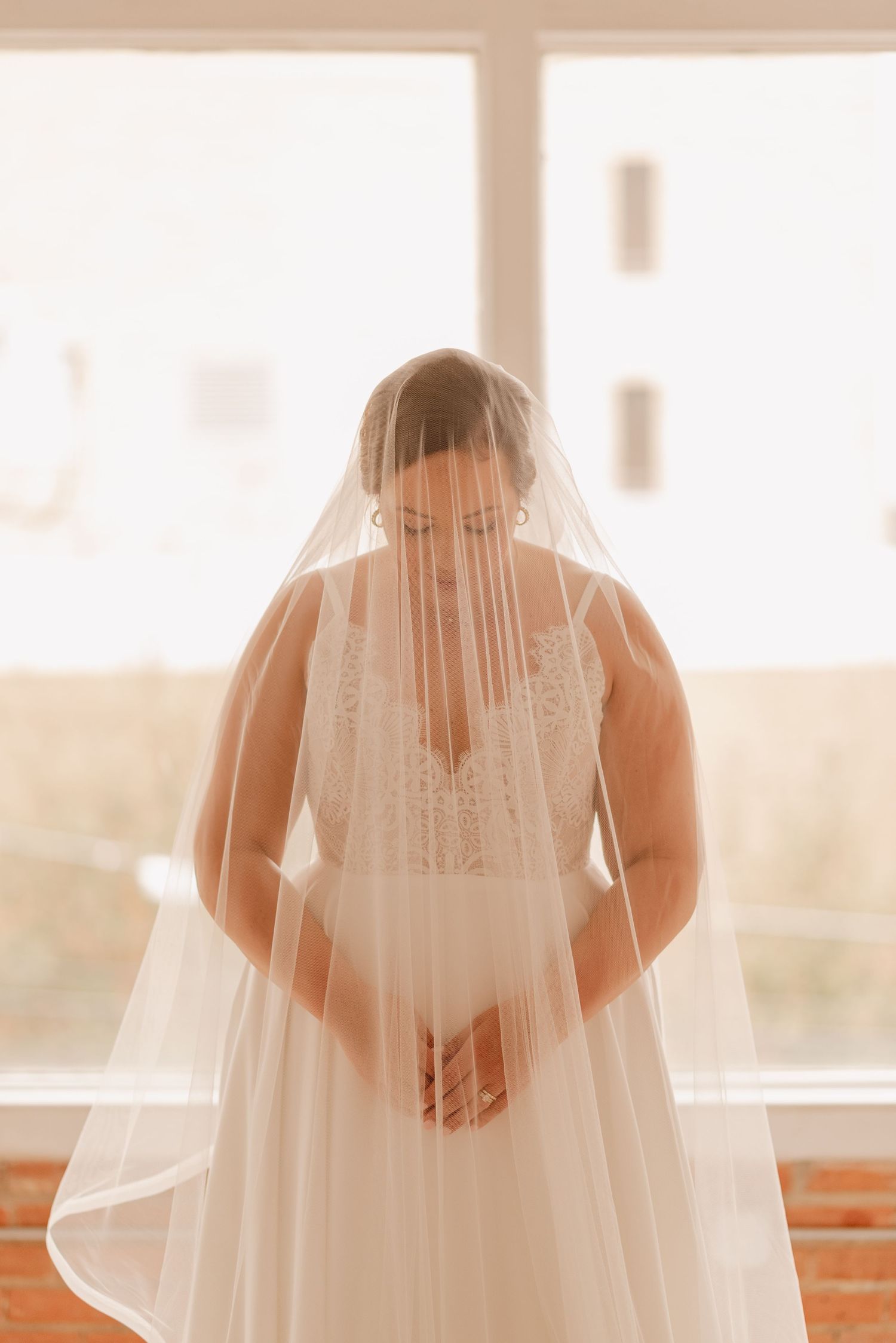 Silhouetted figure in wedding dress with flowing veil backlit by bright windows.