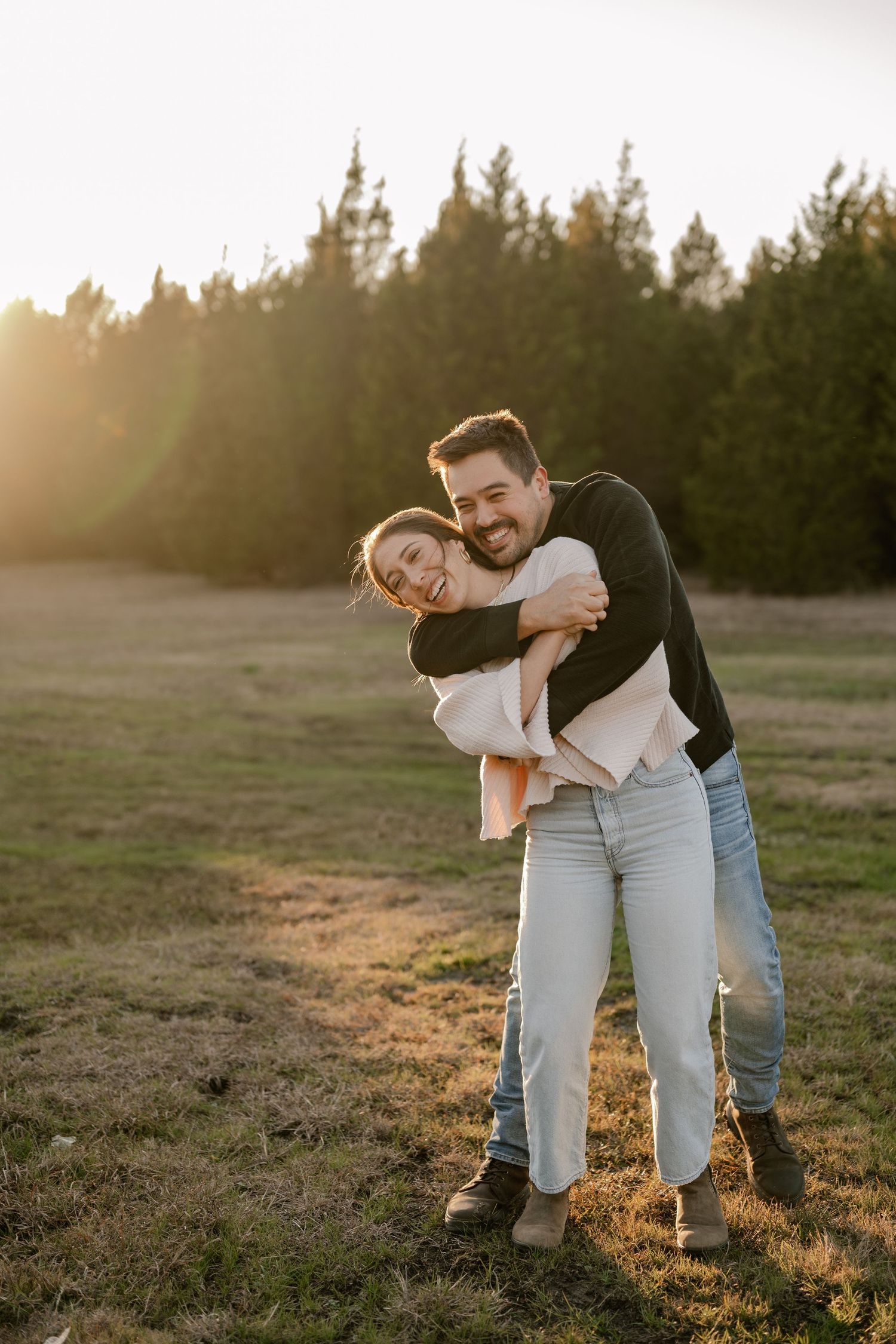 Couple playfully hugging in a larger evergreen tree lined field at golden hour for their couple's session in Sunnyvale, Texas