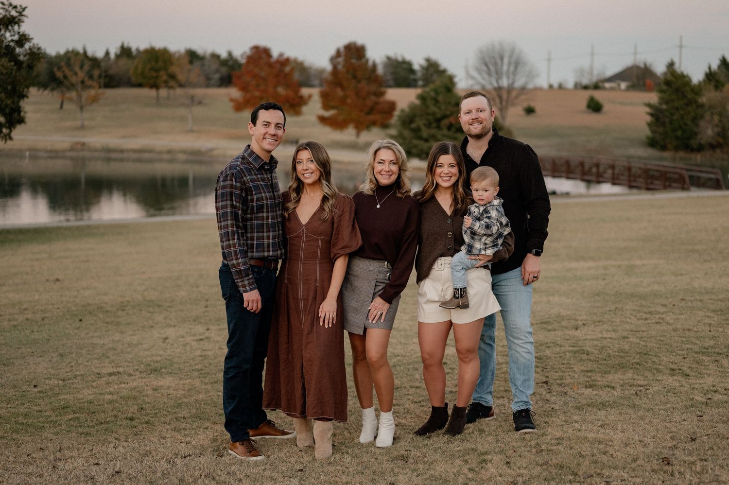Extended family in shades of brown standing in a large field with a pond and colorful trees for a family session in Rockwall, Texas