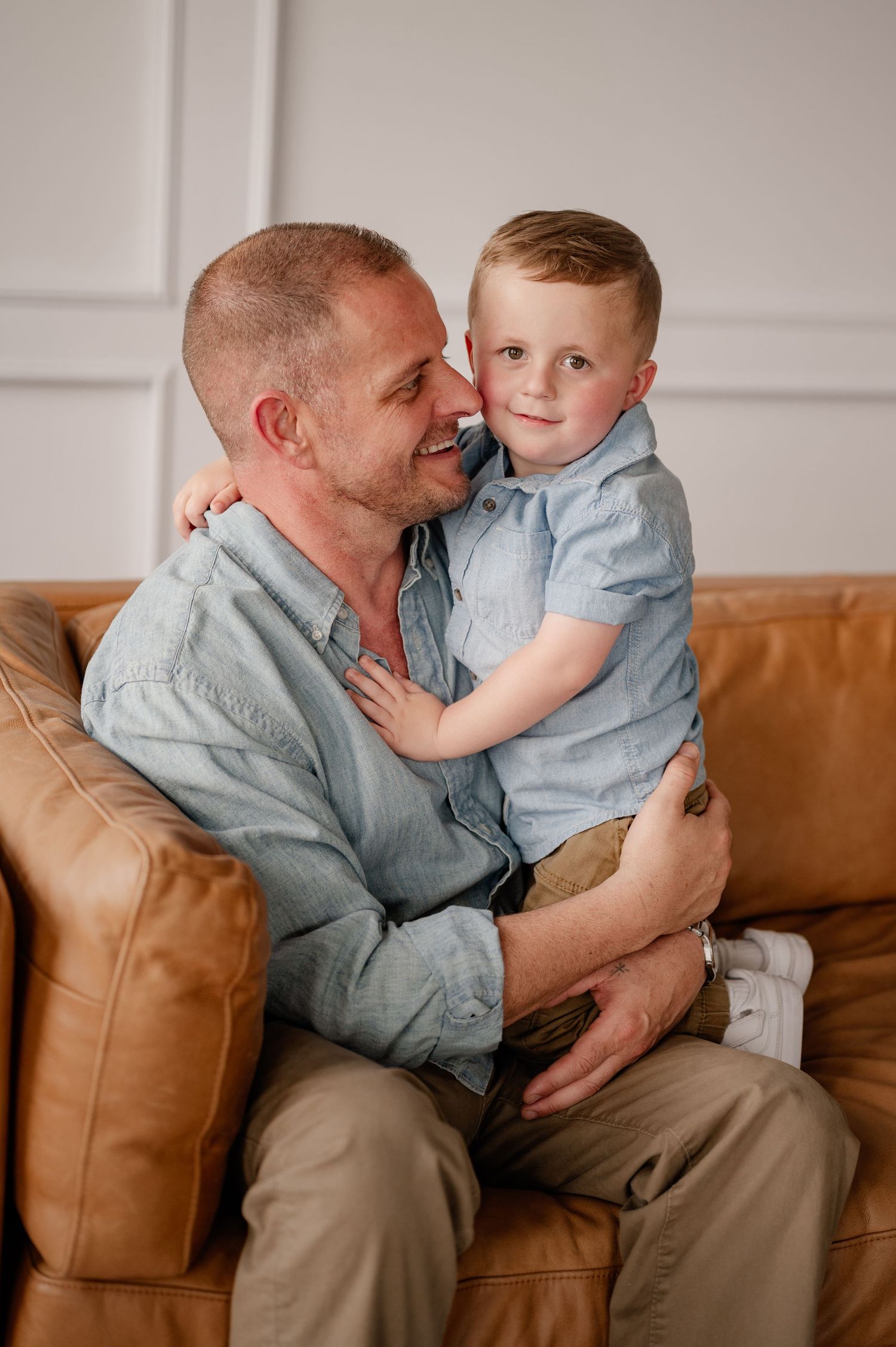 Tender moment shared with dad and son on a brown leather couch in a bright, minimalist studio in Dallas, Texas