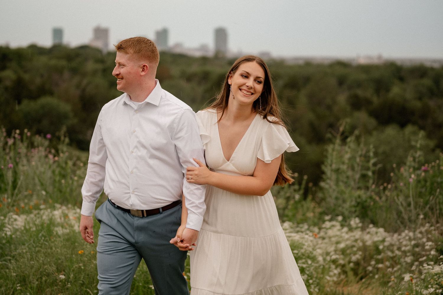 Couple walking hand-in-hand through wildflower field with city skyline backdrop during outdoor engagement photo session.