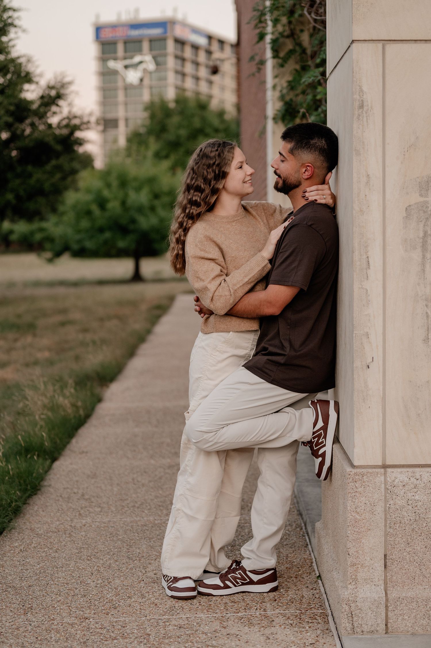 Couple embracing against a stone wall outside of Bush Library on the SMU campus with an urban park background.