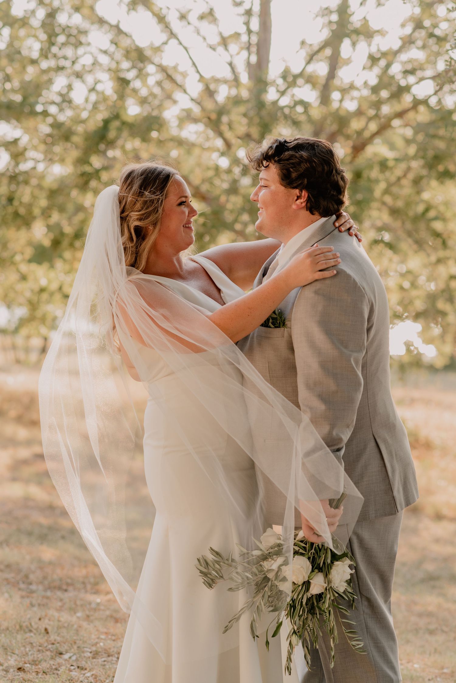Joyful wedding couple embracing outdoors with flowing veil in golden sunlight.