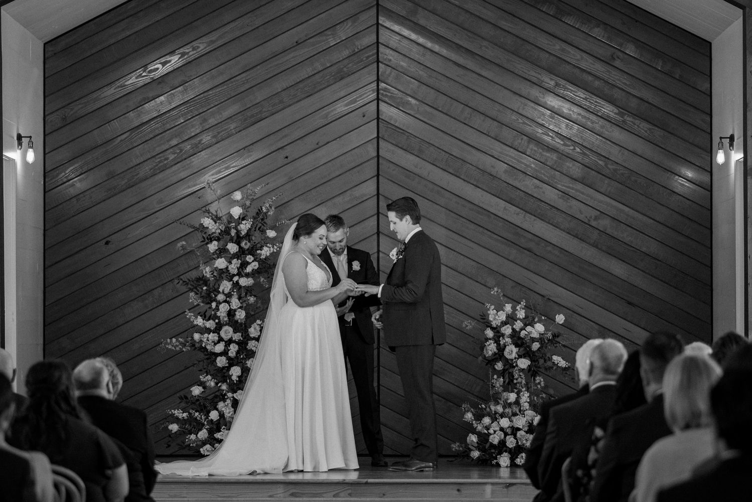 Black and white photo of a wedding ceremony in a wooden chapel with guests seated and beautiful floral decor.