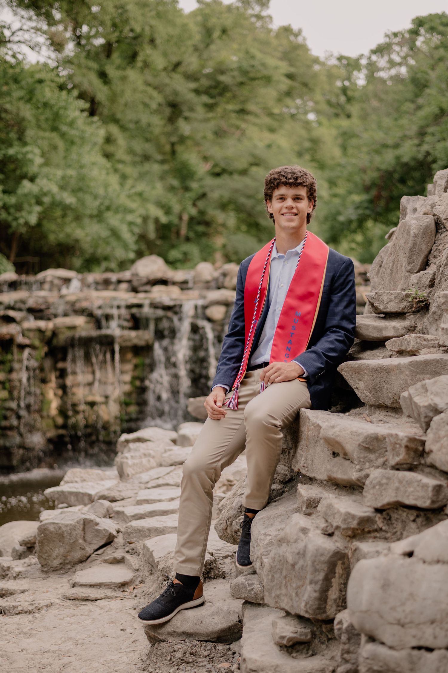 Senior male sitting on large rocks in front of a waterfall at Prairie Creek Park in Richardson, TX for a sunner senior photo session