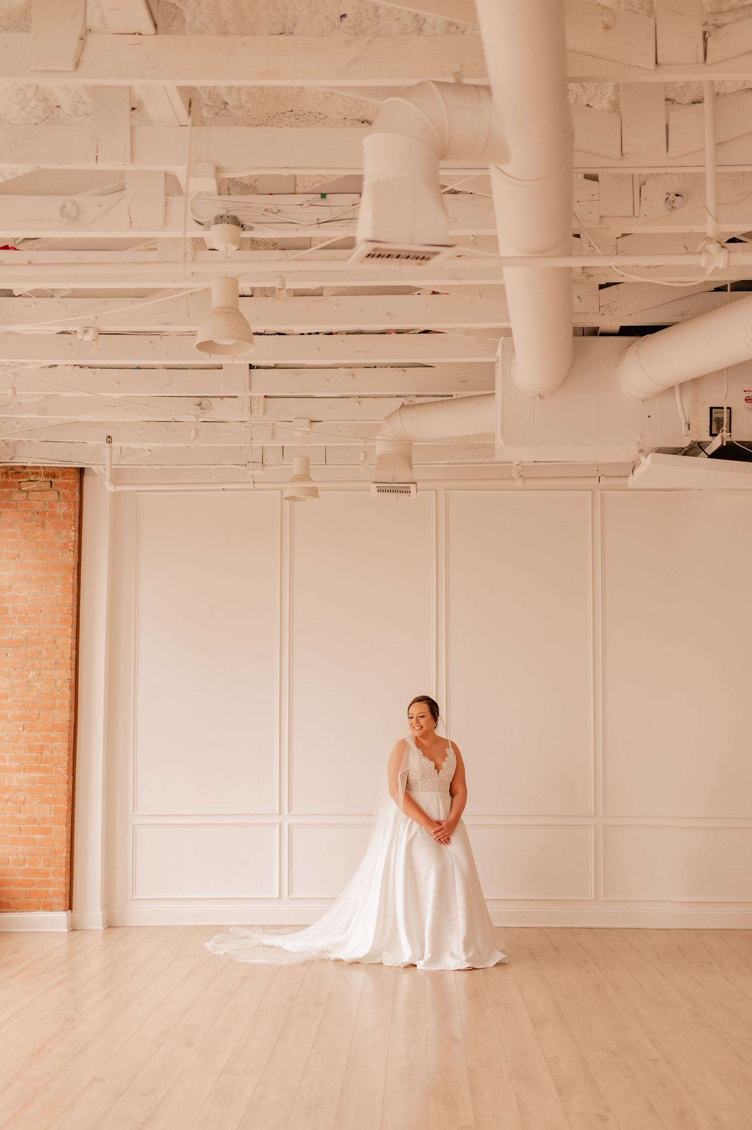 A bride in an elegant white gown stands gracefully in a bright, industrial-style room with exposed ceiling and brick walls.