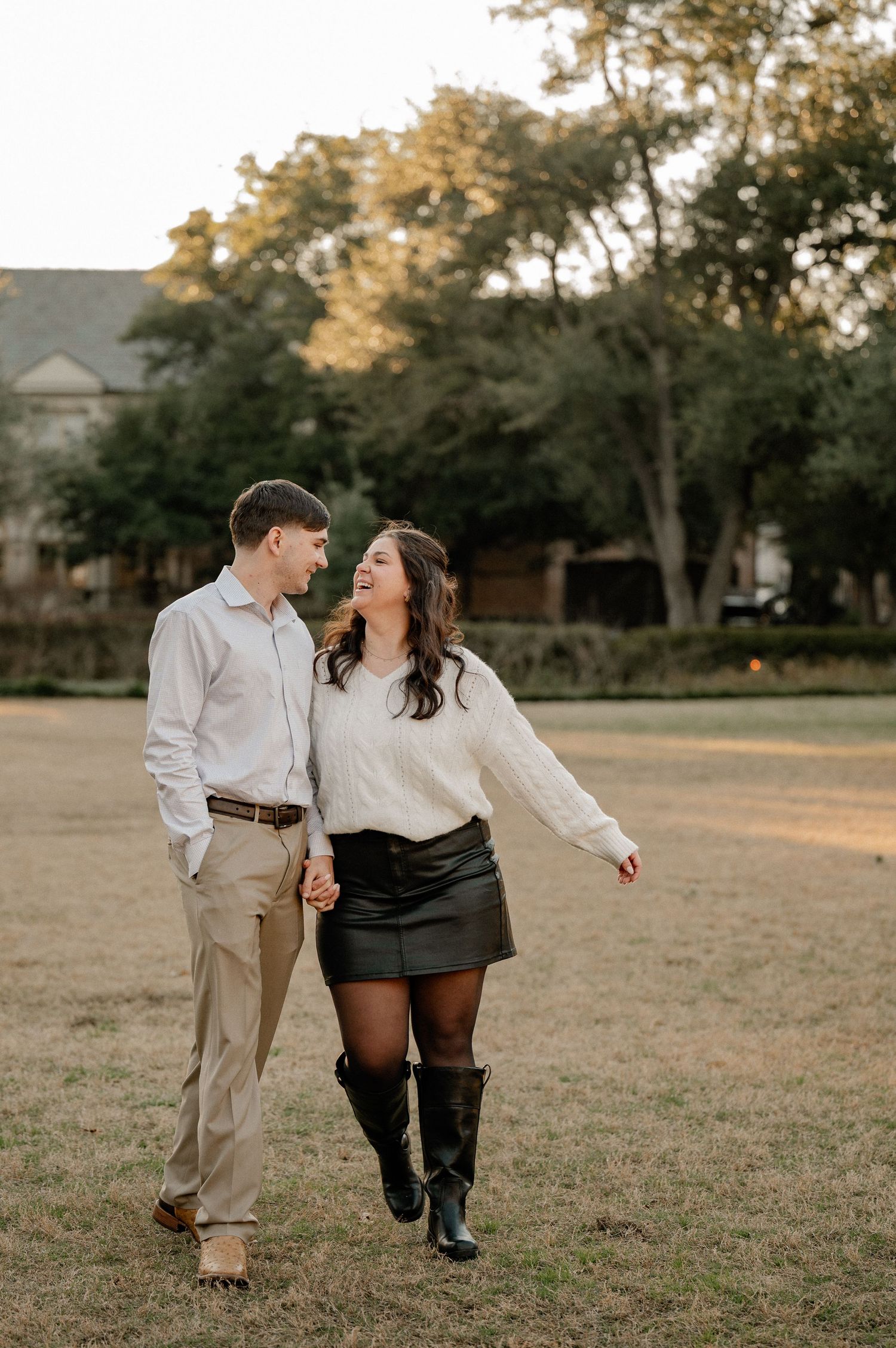 A couple in Highland Park, TX walks hand-in-hand through a grassy park, smiling at each other, dressed in casual autumn outfits.
