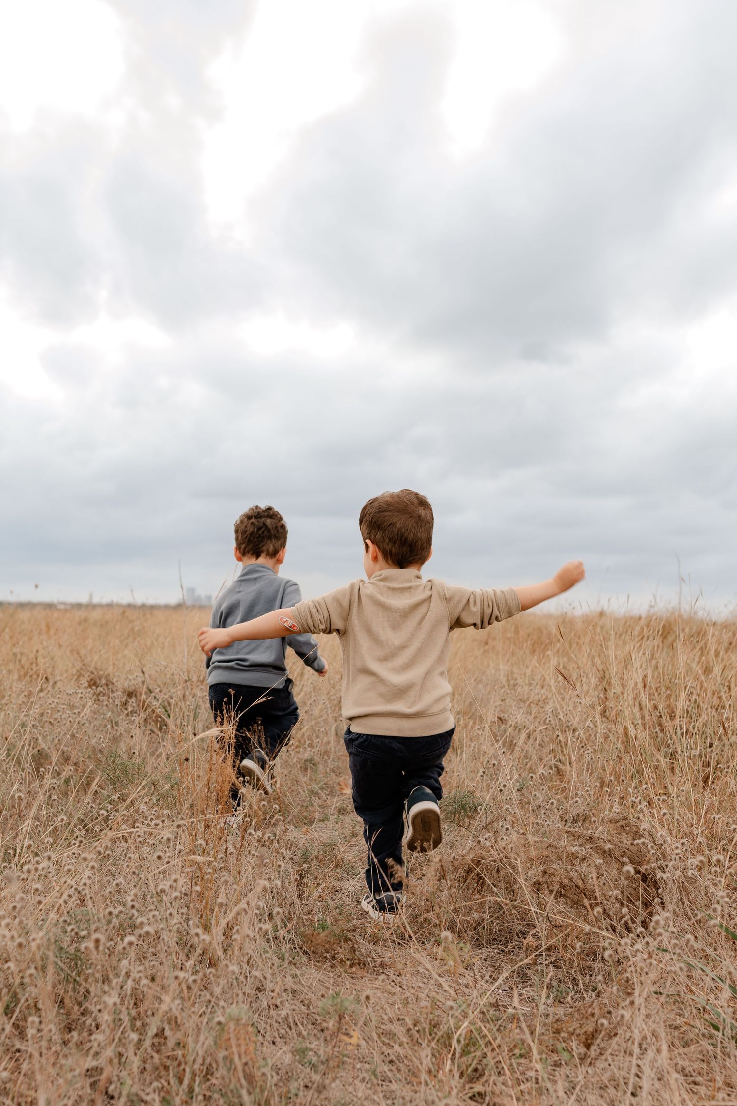 Two boys playfully running away from the camera with airplane arms through tall grass for a fall family photo session