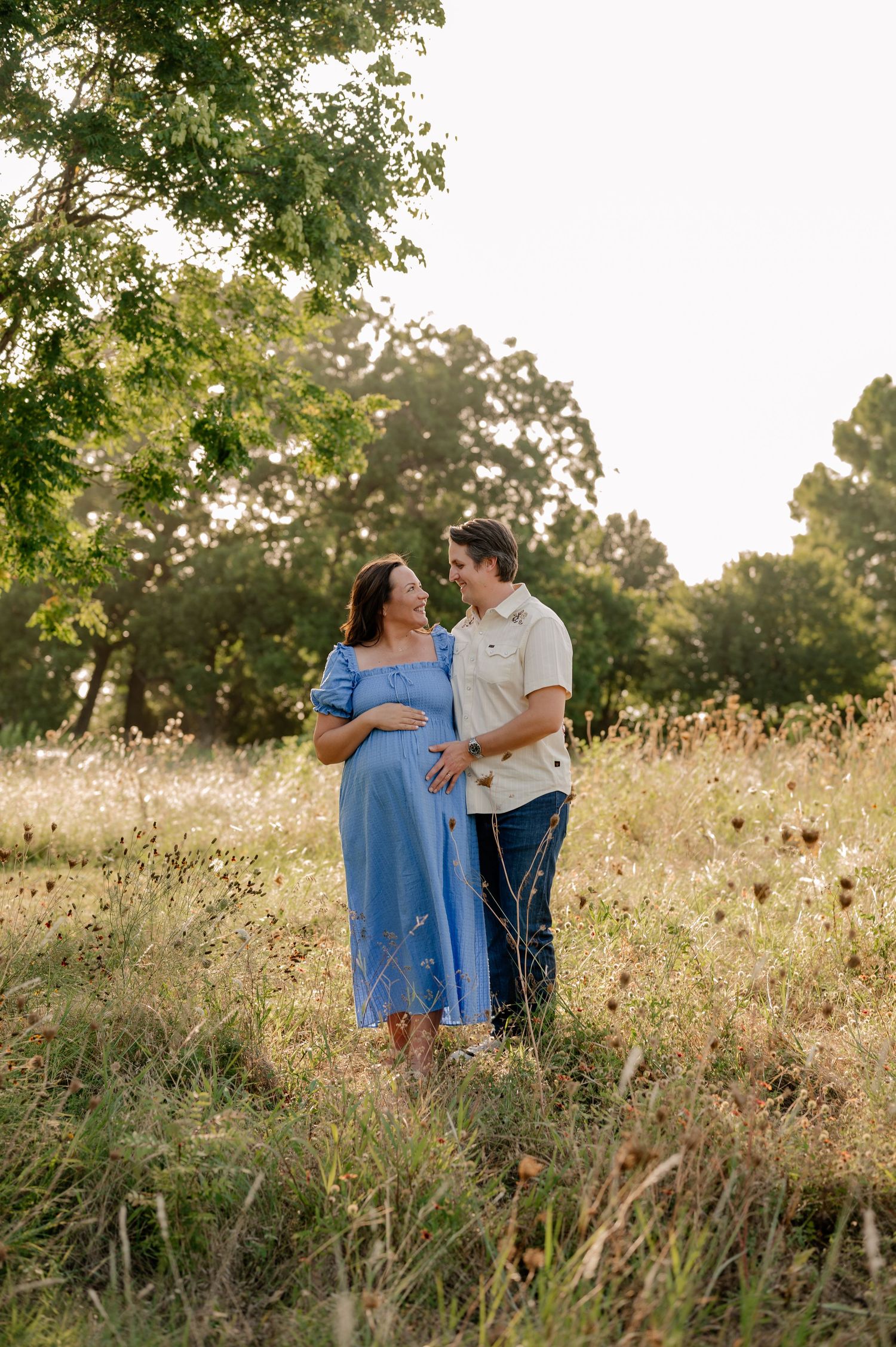Candid photo of couple embracing and hold pregnant belly while in a large wild grass field in Dallas, Texas for maternity session