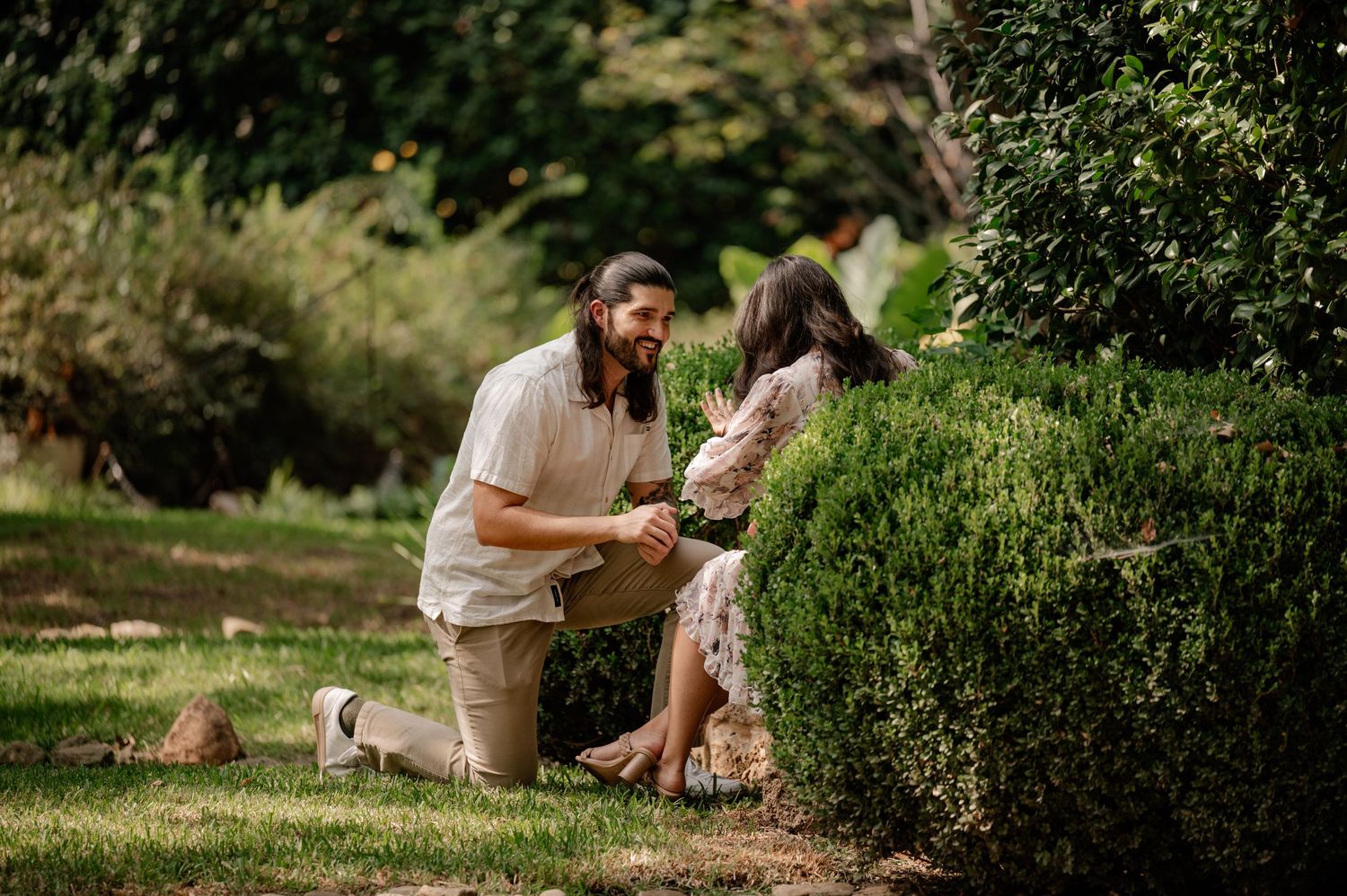 Man down on one knee surrounded by green trees and flowers during a proposal couple's session at the Dallas Arboretum in East Dallas