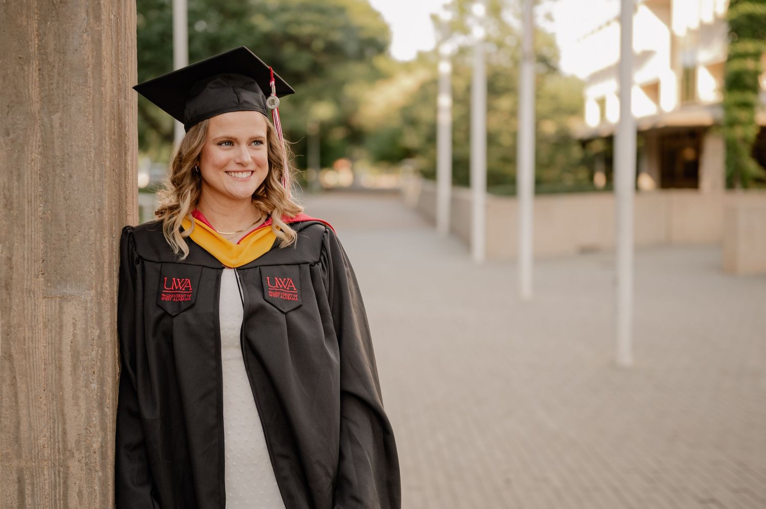 College senior in her cap in gown resting on a pillar for a summer senior portrait session in Richardson, Texas