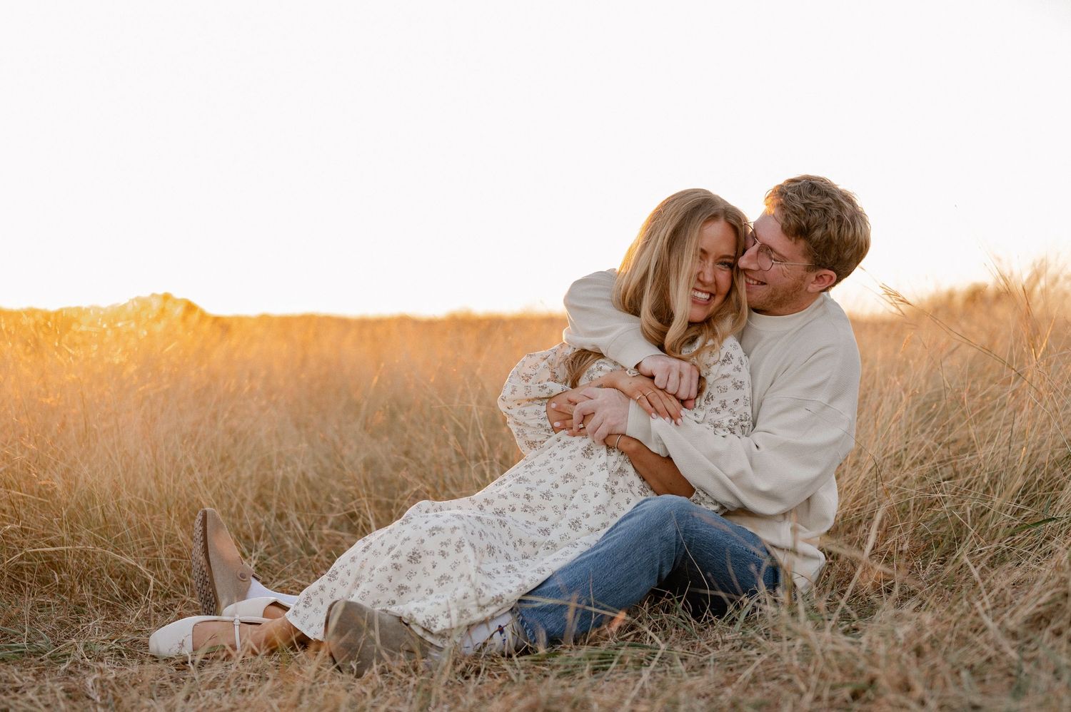 Couple sitting and playfully cuddled up in a tall grass autumn field with treelined backdrop during engagement photo session.