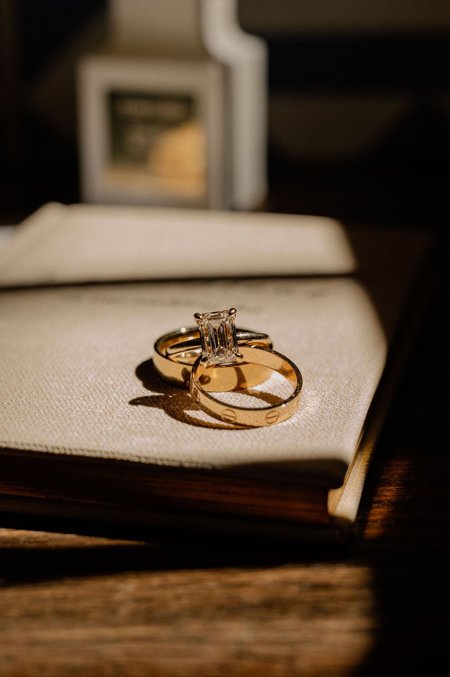 Gold engagement ring with emerald-cut diamond stacked on wedding band resting on an open book in warm dramatic light