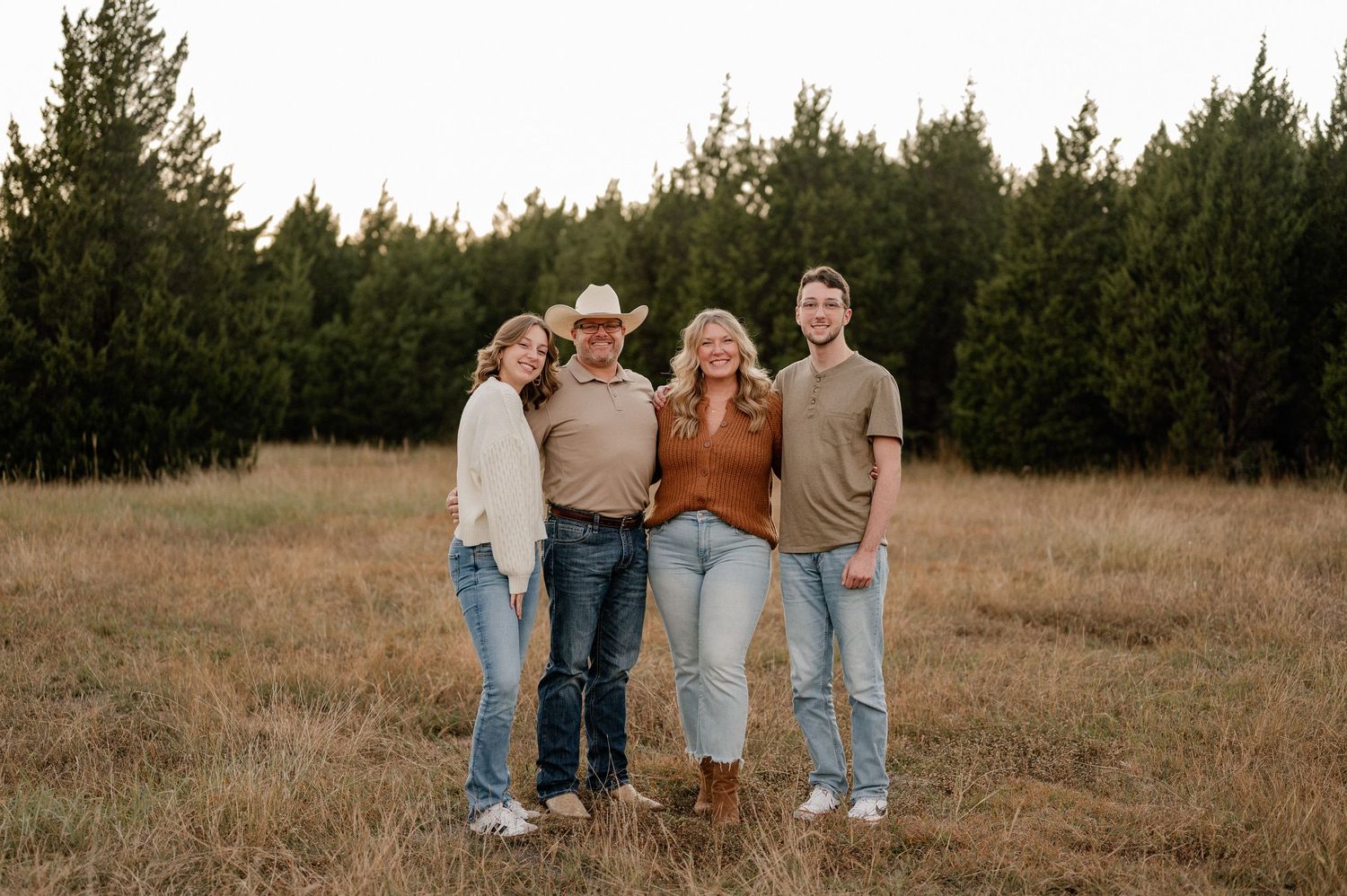 Family of four posing for fall family photos in a large tree lined field in warm colors during blue hour