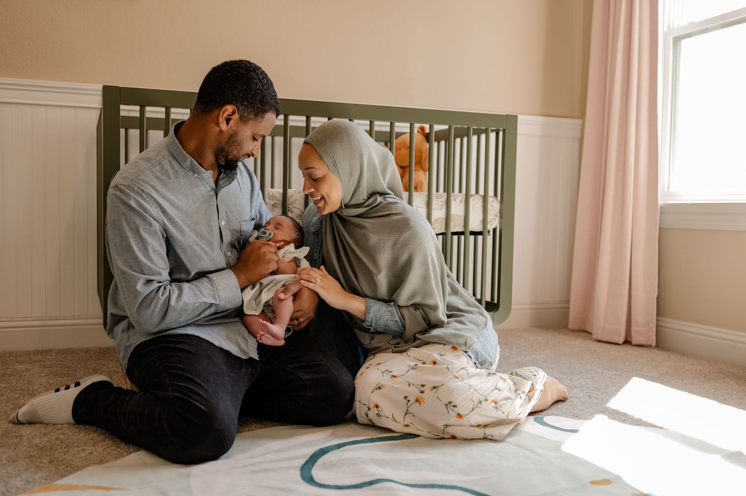 Husband holding newborn while he and wife staring at her in their bright warm nursery for an at-home newborn photo session