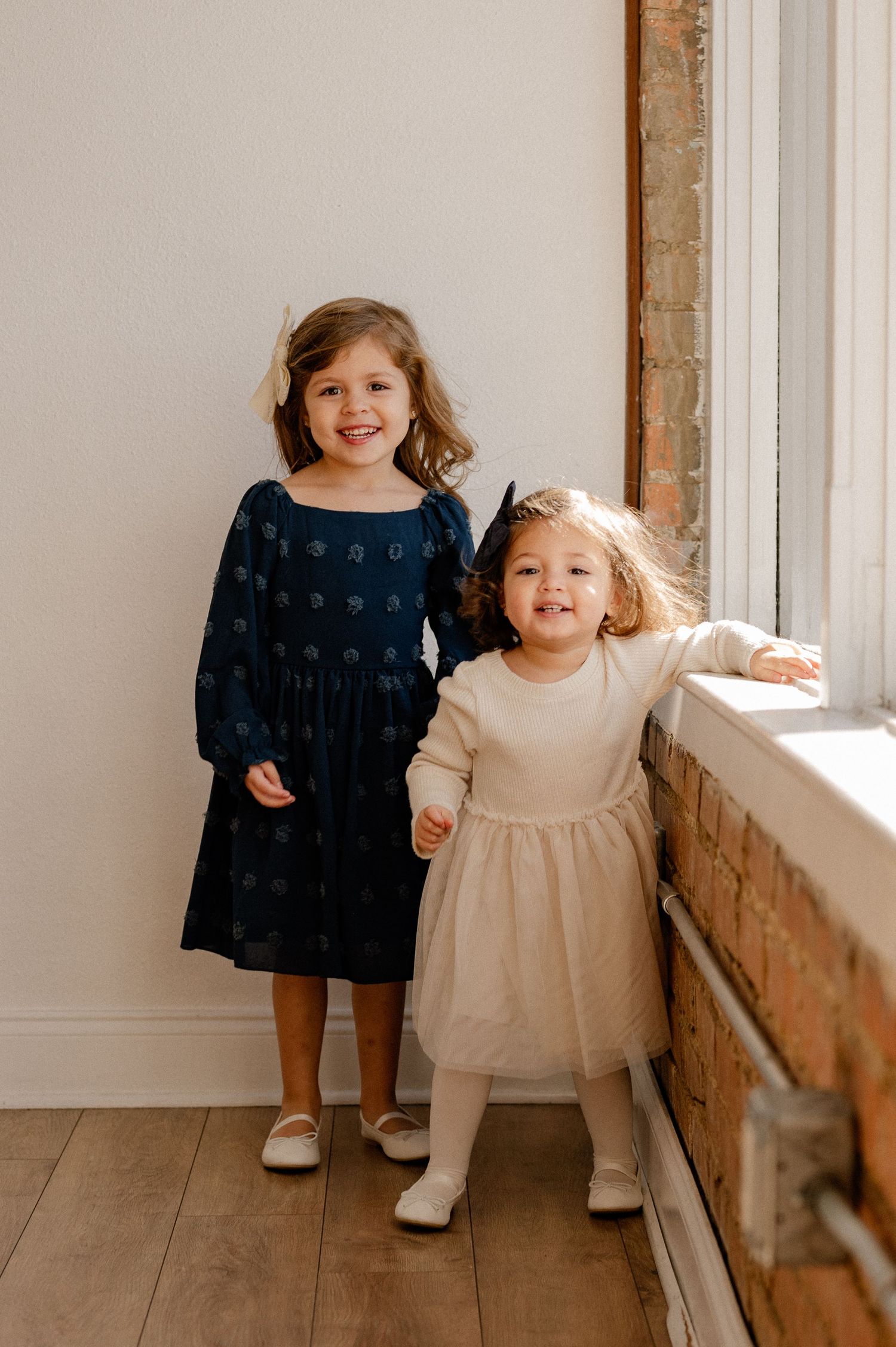 Sisters smiling at the camera in a bright brick room at The Lumen Room Studio for a family photo session in Dallas, Texas