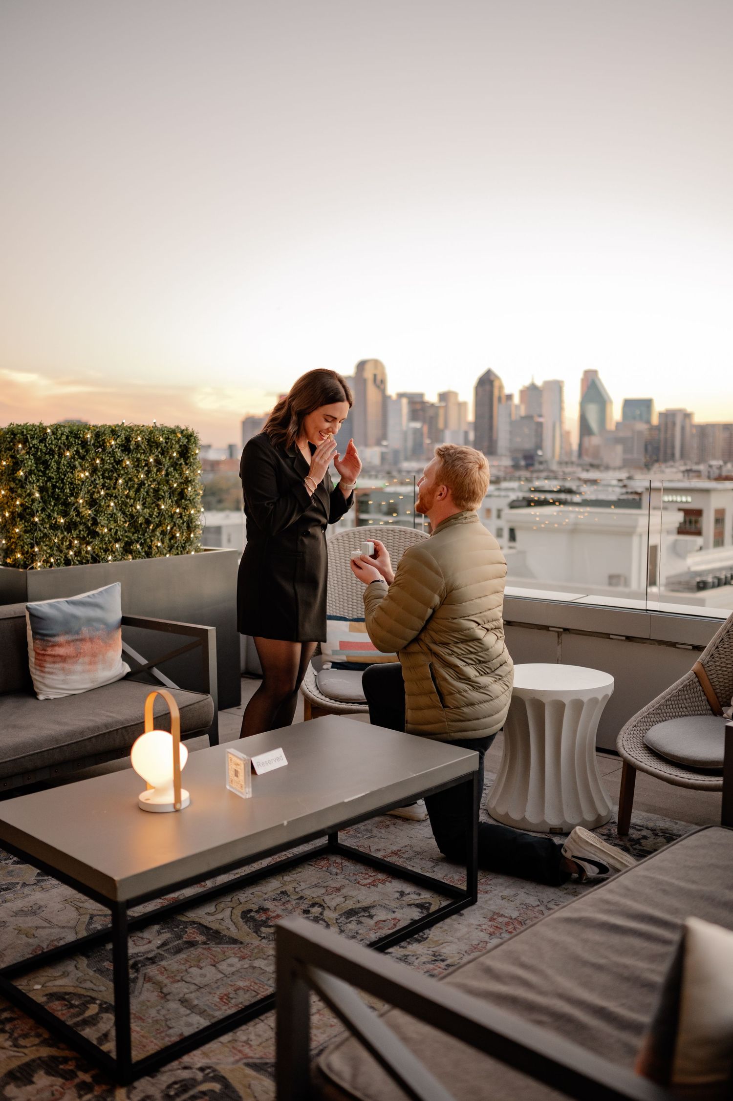 Man on one knee proposing to his girlfriend at a uptown Dallas rooftop bar with the Dallas skyline at dusk in the background