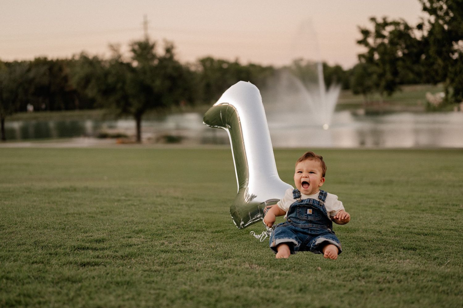 Memento session of child sitting next to a 1 balloon in a large field with a pond and water feature in the background in Rockwall, Texas