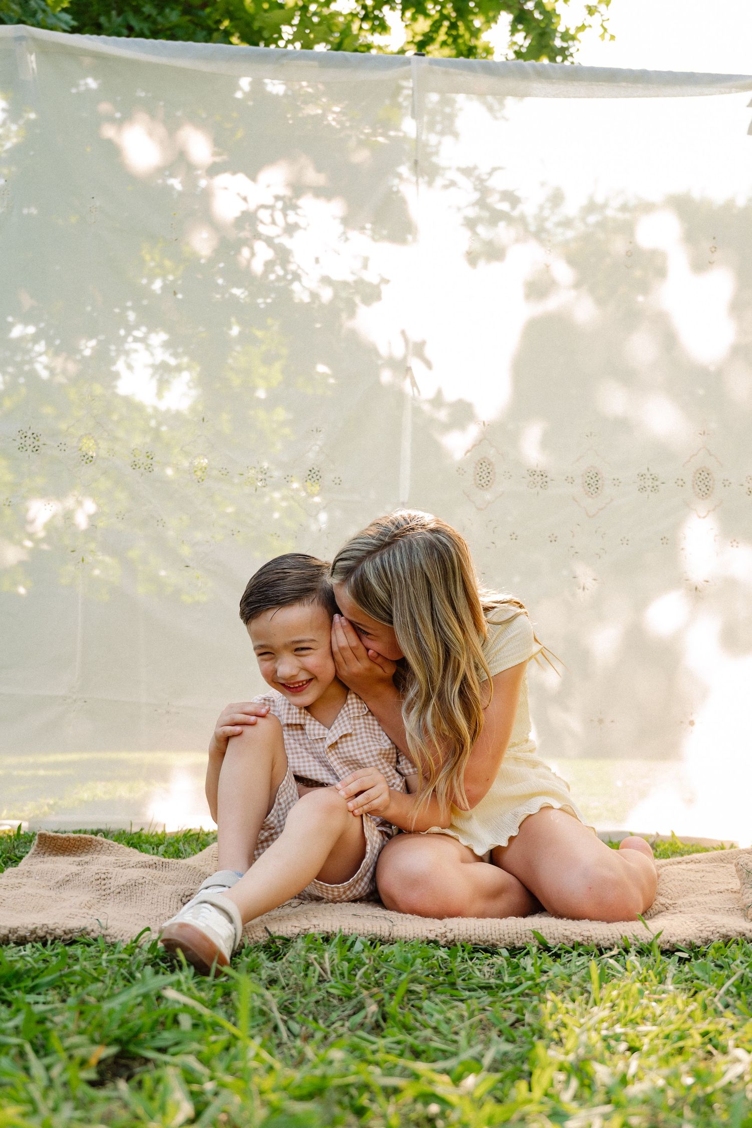 Two little siblings whispering and giggling at an at-home summer backyard family photo session in Dallas, Texas
