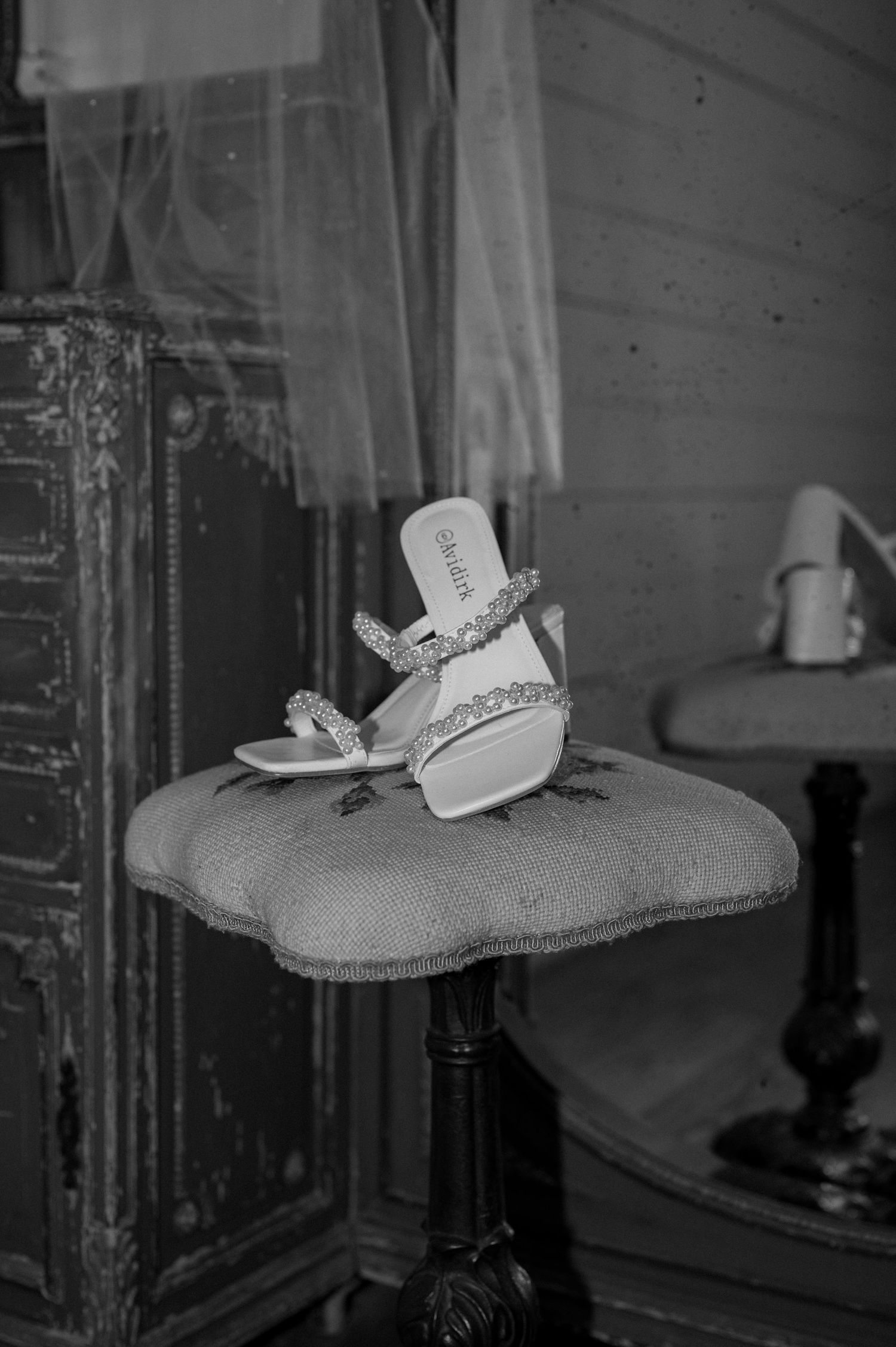 Black and white photo of embellished bridal shoes displayed on a vintage upholstered stool in an elegant room.