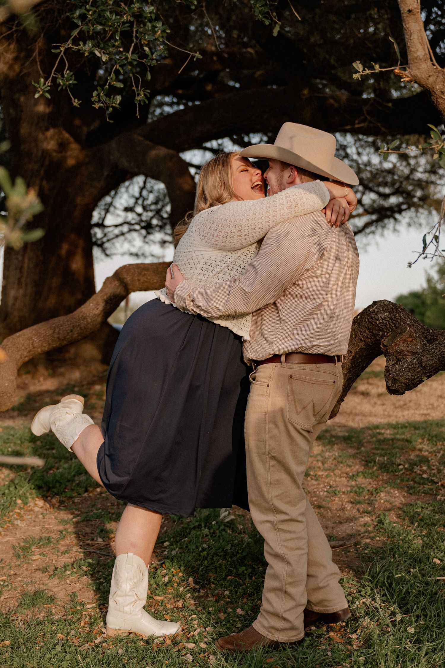 Couple sharing a romantic kiss outdoors, woman in white cowboy boots and dark skirt, man in cowboy hat, under a large tree.