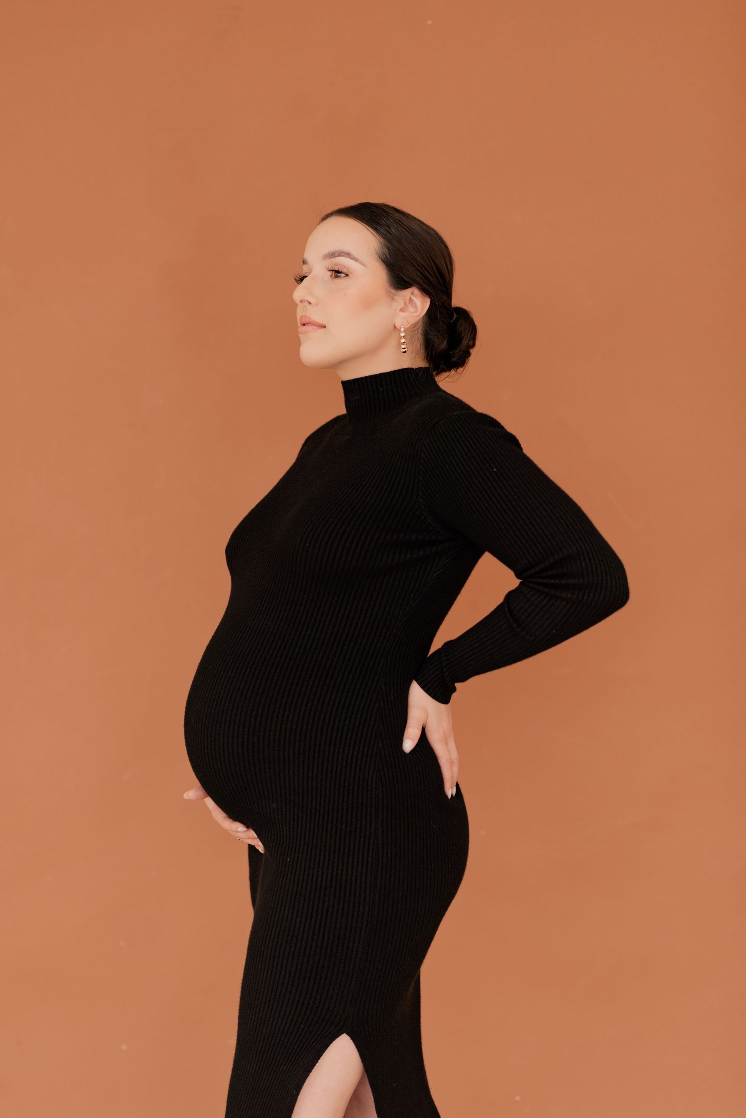 Pregnant woman in sleek black dress holding her belly in front of a burnt orange backdrop for a studio maternity photo session in Dallas, TX
