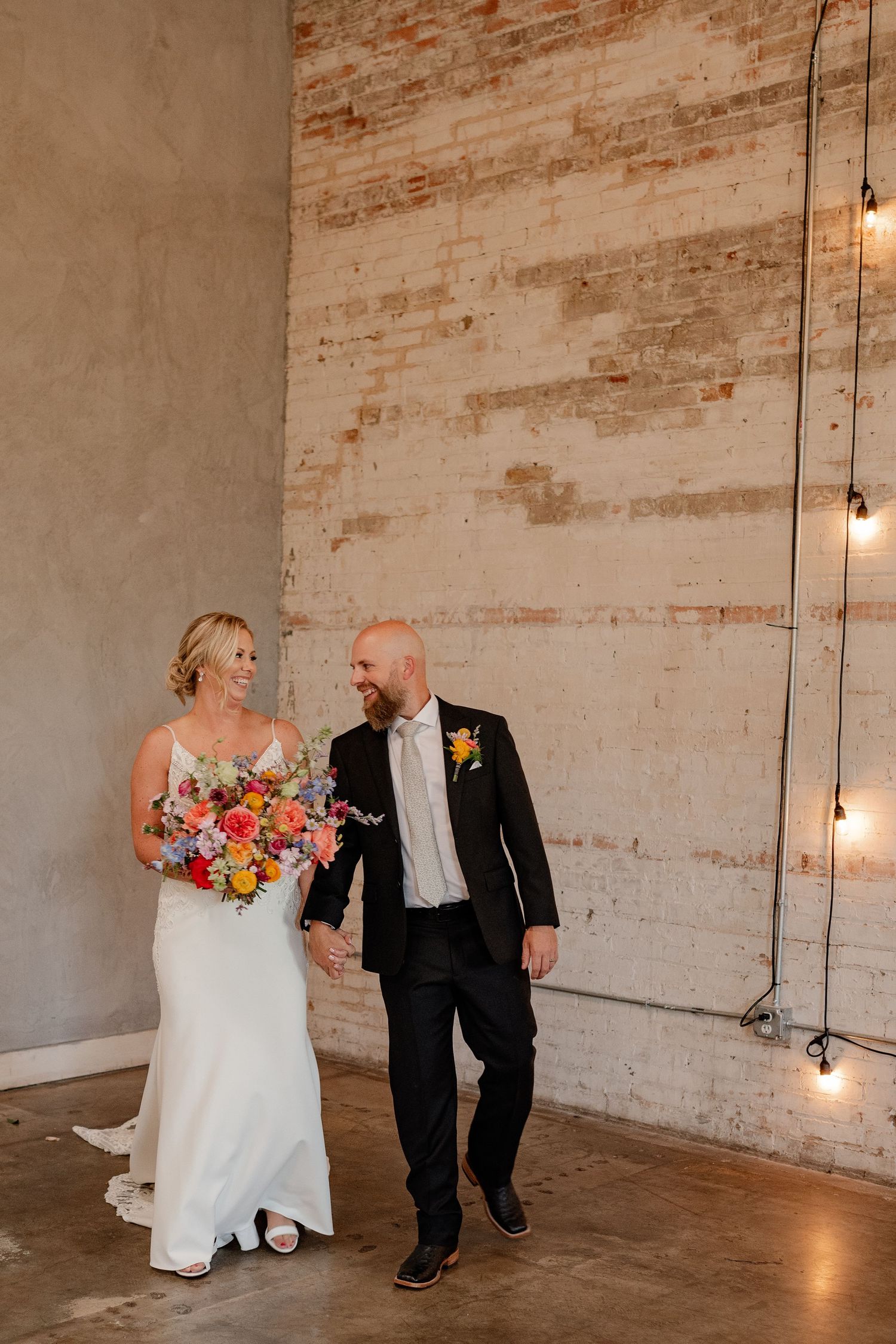 Bride and Groom walking and laughing during wedding portraits in front of industrial brick-walled venue in Deep Ellum area of Dallas, Texas