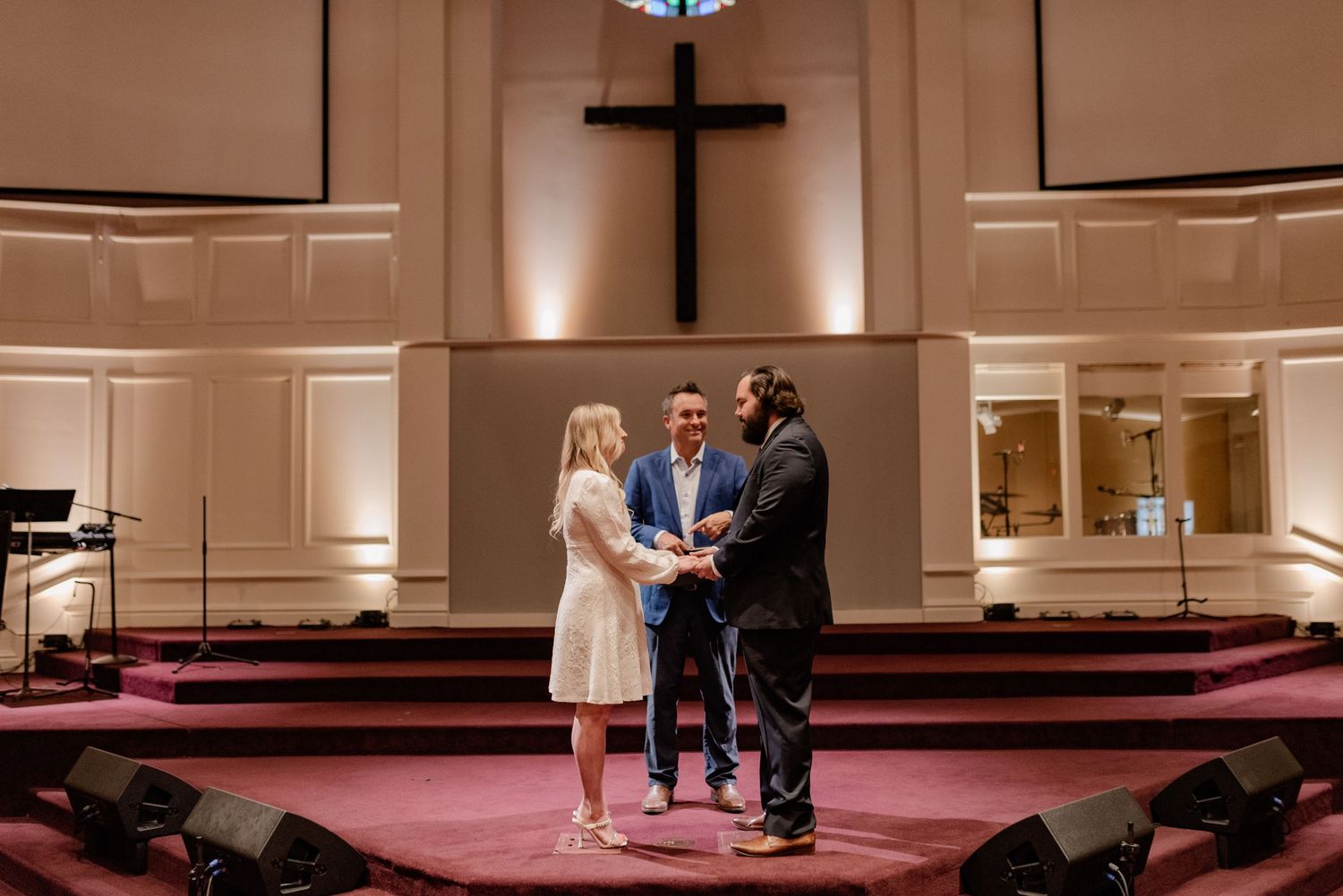 Intimate church wedding ceremony taking place in front of a large wooden cross on stage with warm lighting.