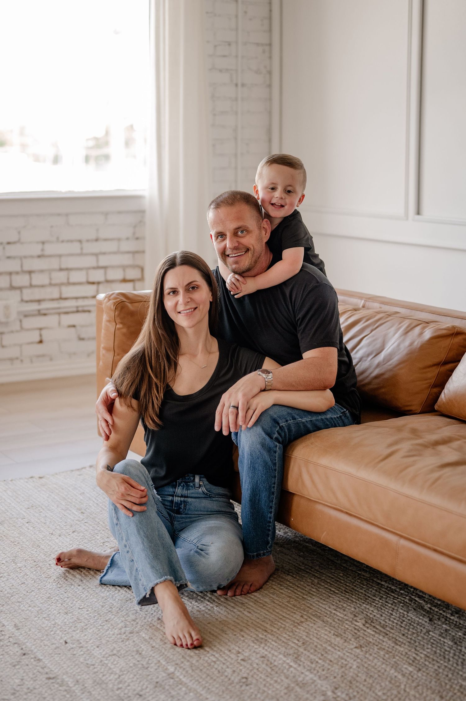 Family of 3 sitting on a couch smiling in a white brick walled studio at The Lumen Room in Dallas TX for an indoor family photo session