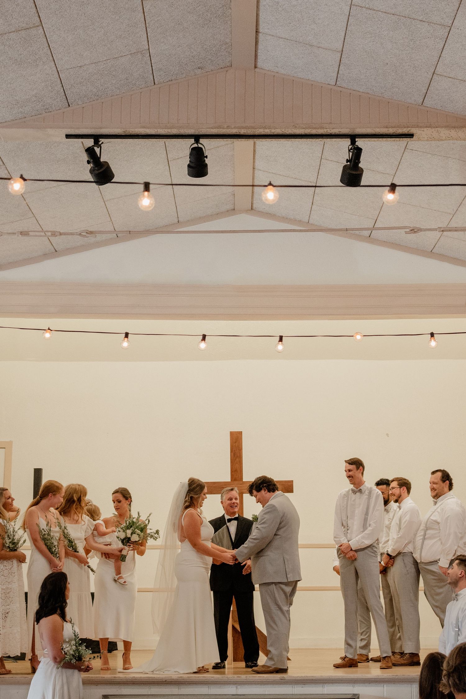 Wedding ceremony guests gather in a bright indoor venue with string lights and white walls.