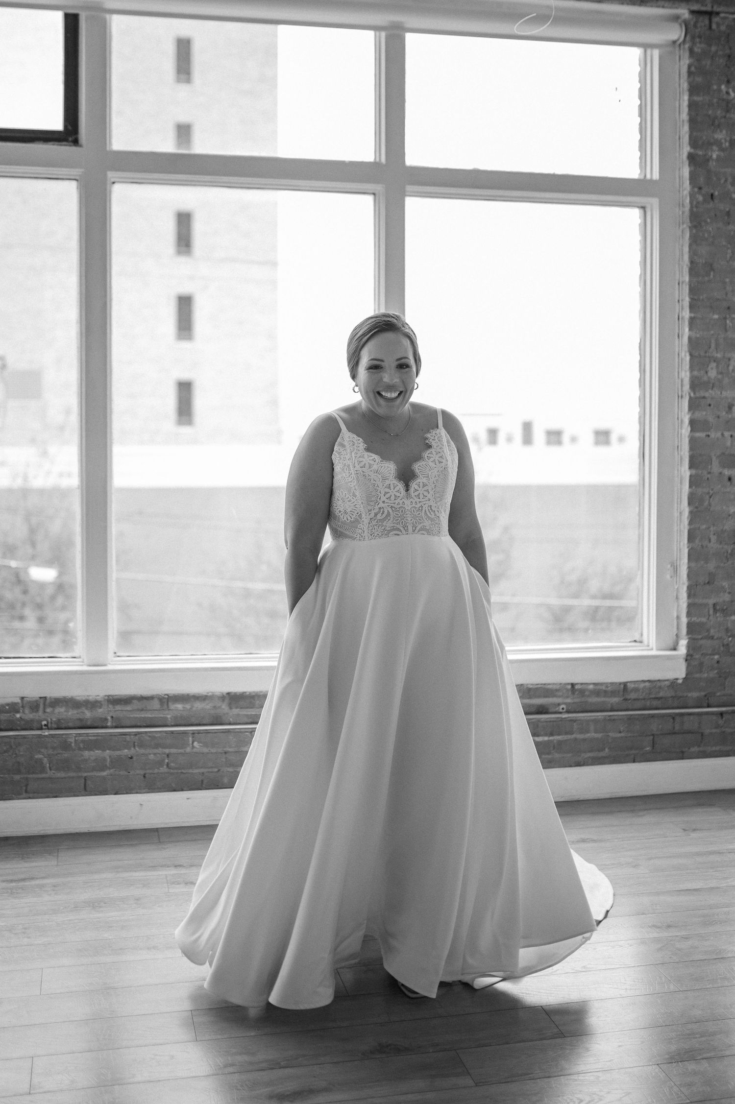 Black and white portrait of bride in full-skirted wedding gown standing by large windows.