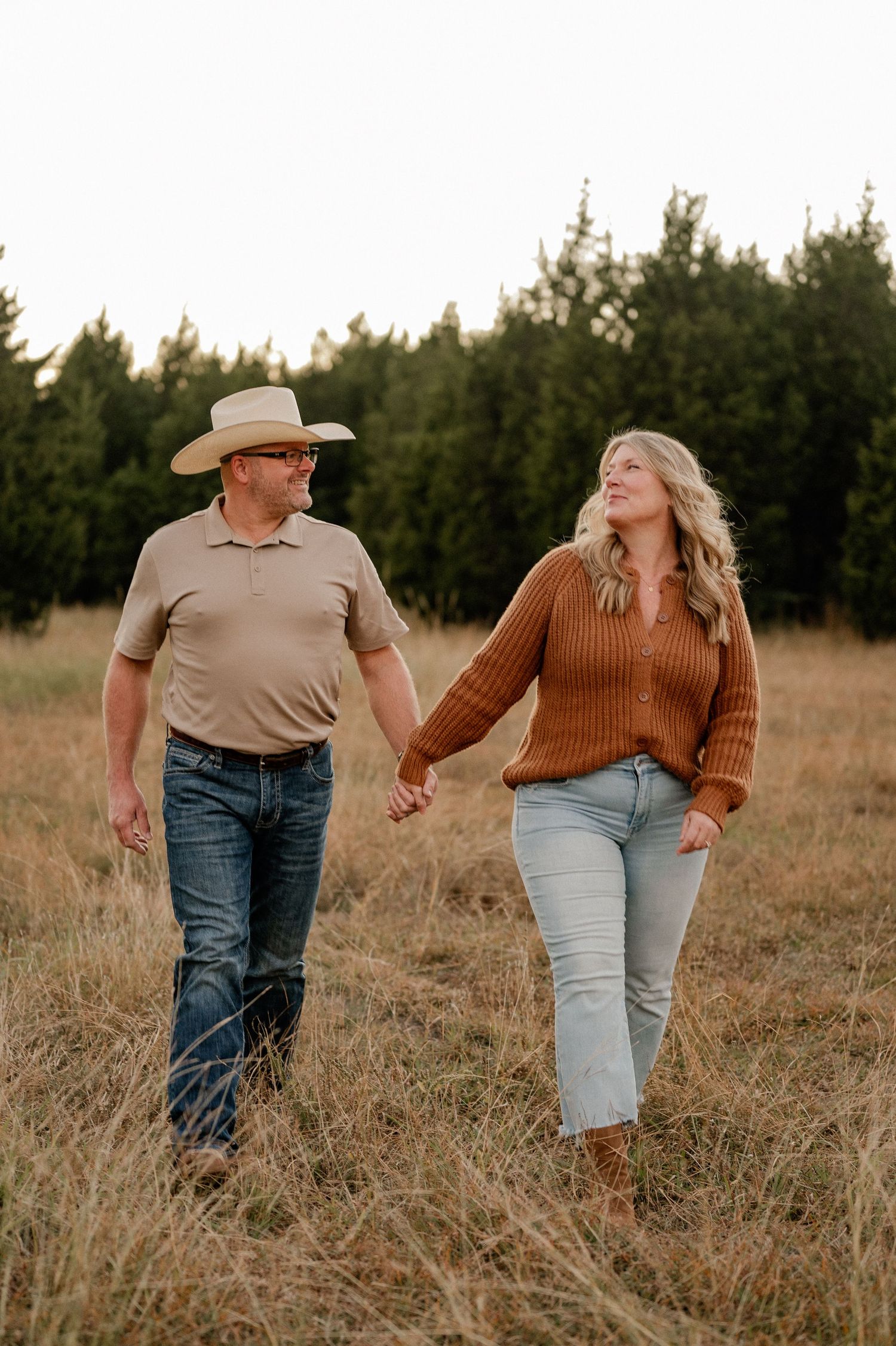 Couple walking hand-in-hand through tall grass autumn field with treelined backdrop during a family photo session.