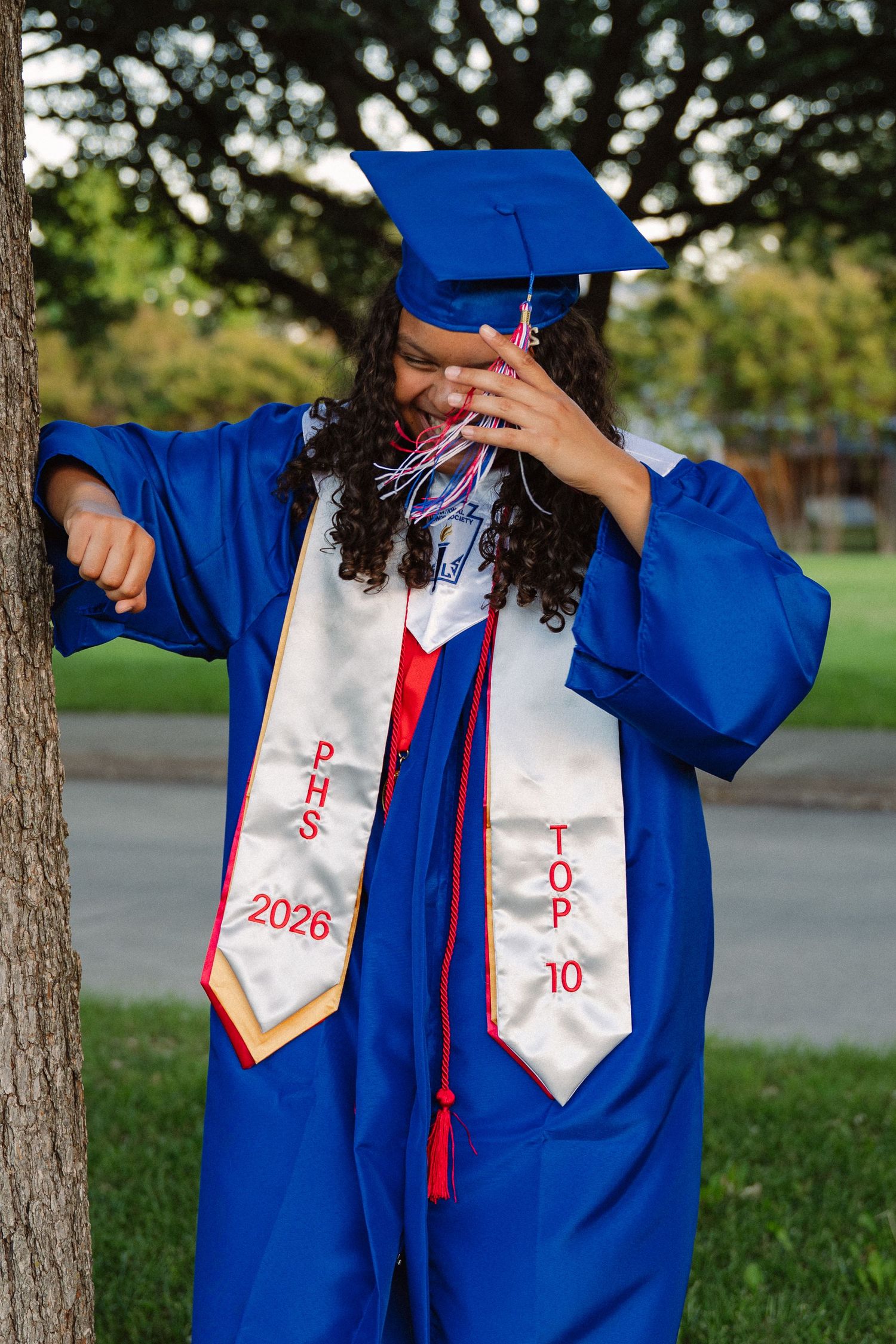 Senior in her cap, gown, and awards playing with her tassel in a large tree-filled park in Richardson, Texas for a senior photo session