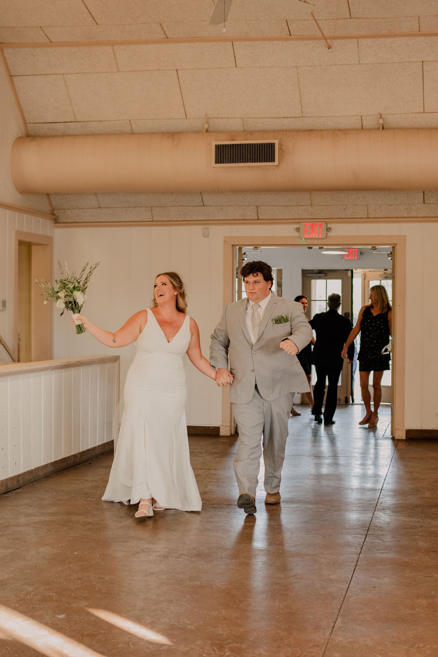 Wedding party dancing in a spacious venue with white walls and sunlight streaming through.