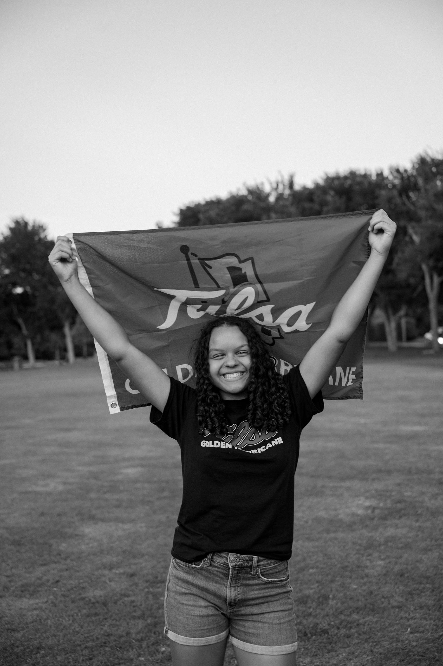 Black and white image of a senior girl holding a college banner sitting in a large field for a Spring senior portrait session