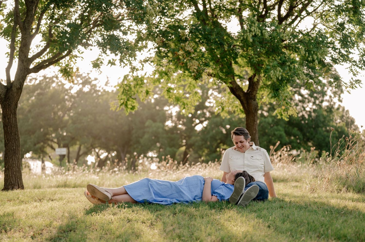 Dreamy candid of a wife laying her head on her husband's lap while holding her pregnant belly under tree shade overlooking White Rock Lake
