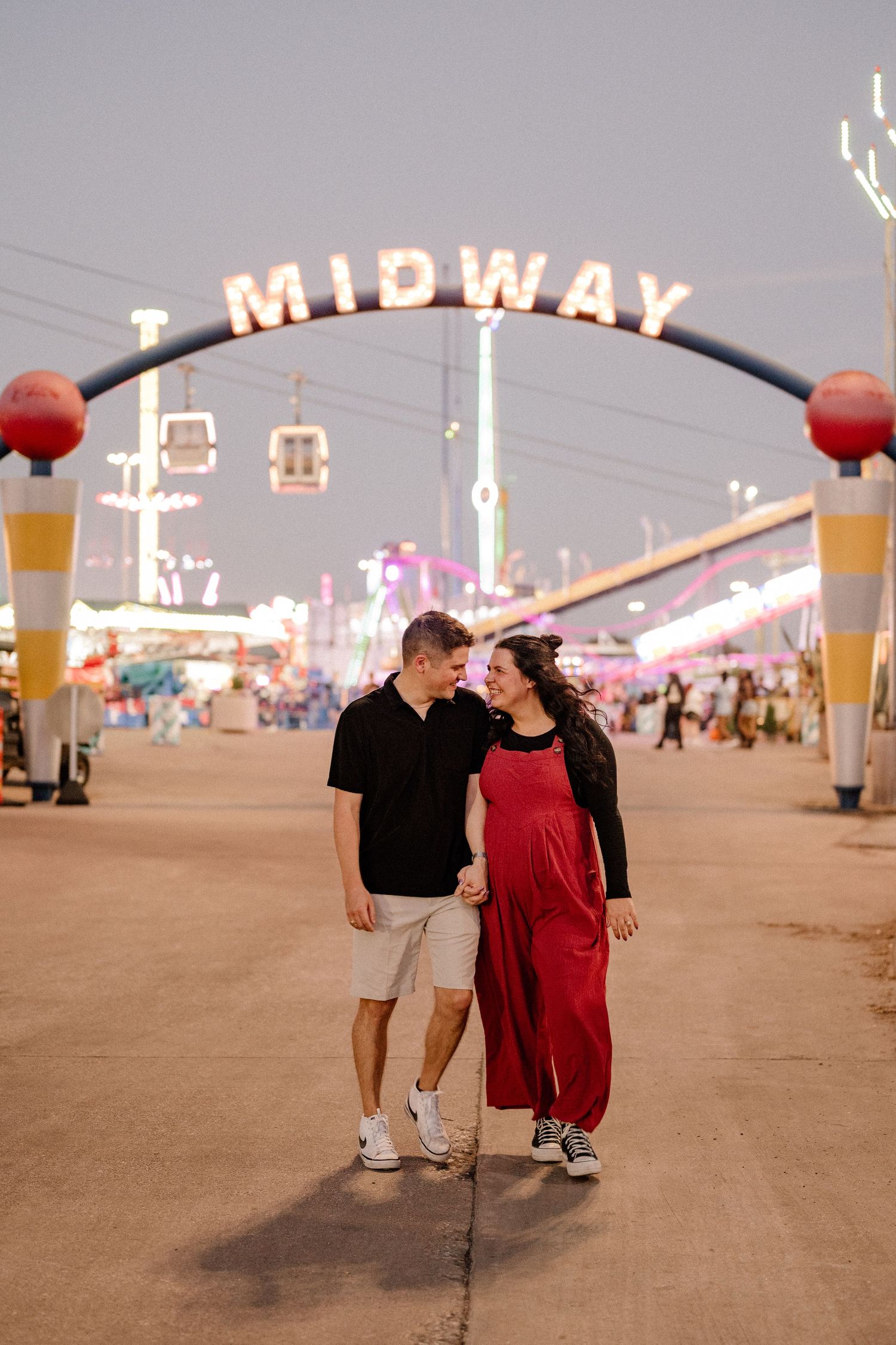 Expecting parents playfully walking through a carnival midway at the State Fair of Texas for a maternity photo shoot