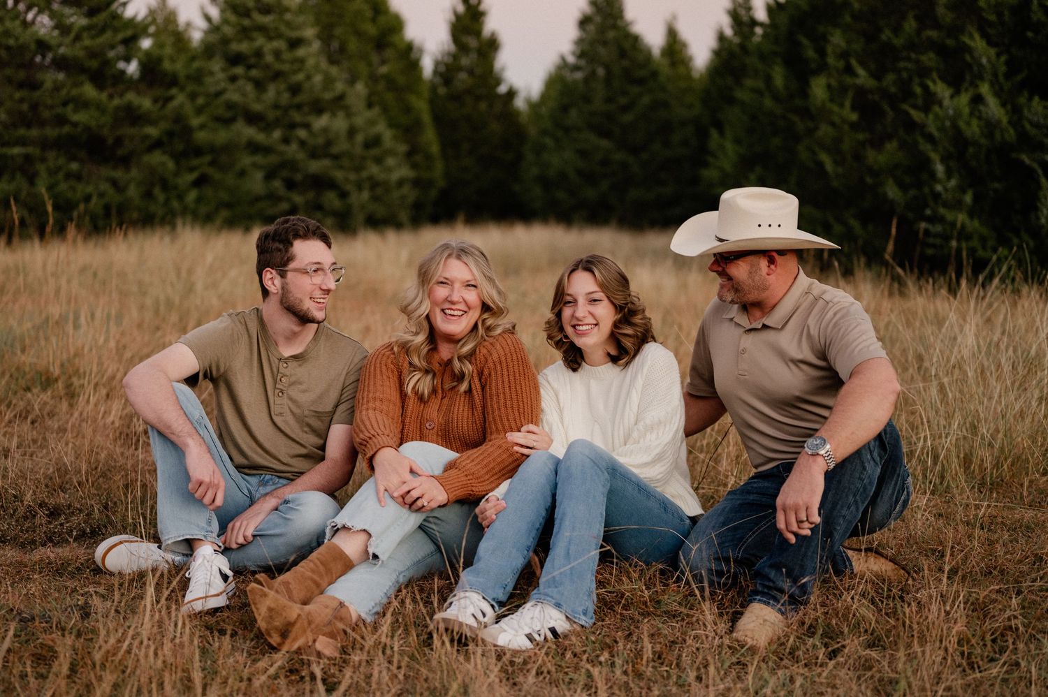 Family of four sitting and laughing candidly for fall family photos in a large tree lined field in warm colors during blue hour