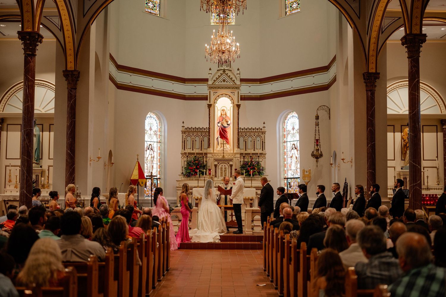 Wide photo of a grandiose Catholic church wedding ceremony with bride, groom, and wedding party lining the front of the sanctuary