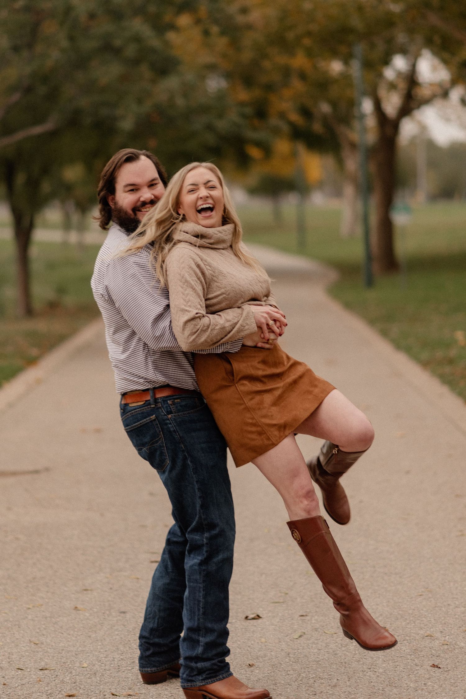 A laughing couple poses playfully on a park path for a fall engagement photo session, wearing fall outfits with boots and jeans.