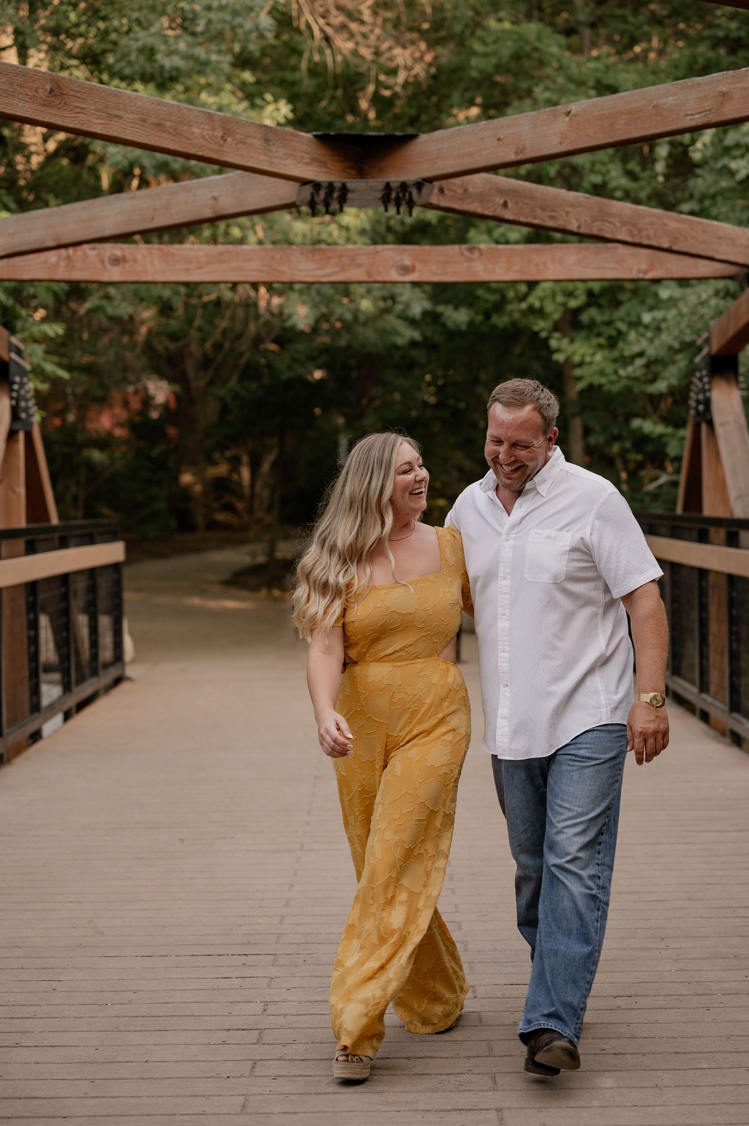 Couple laughing and walking hand-in-hand on a wooden bridge surrounded by lush green trees at Prairie Creek Park in Richardson, Texas